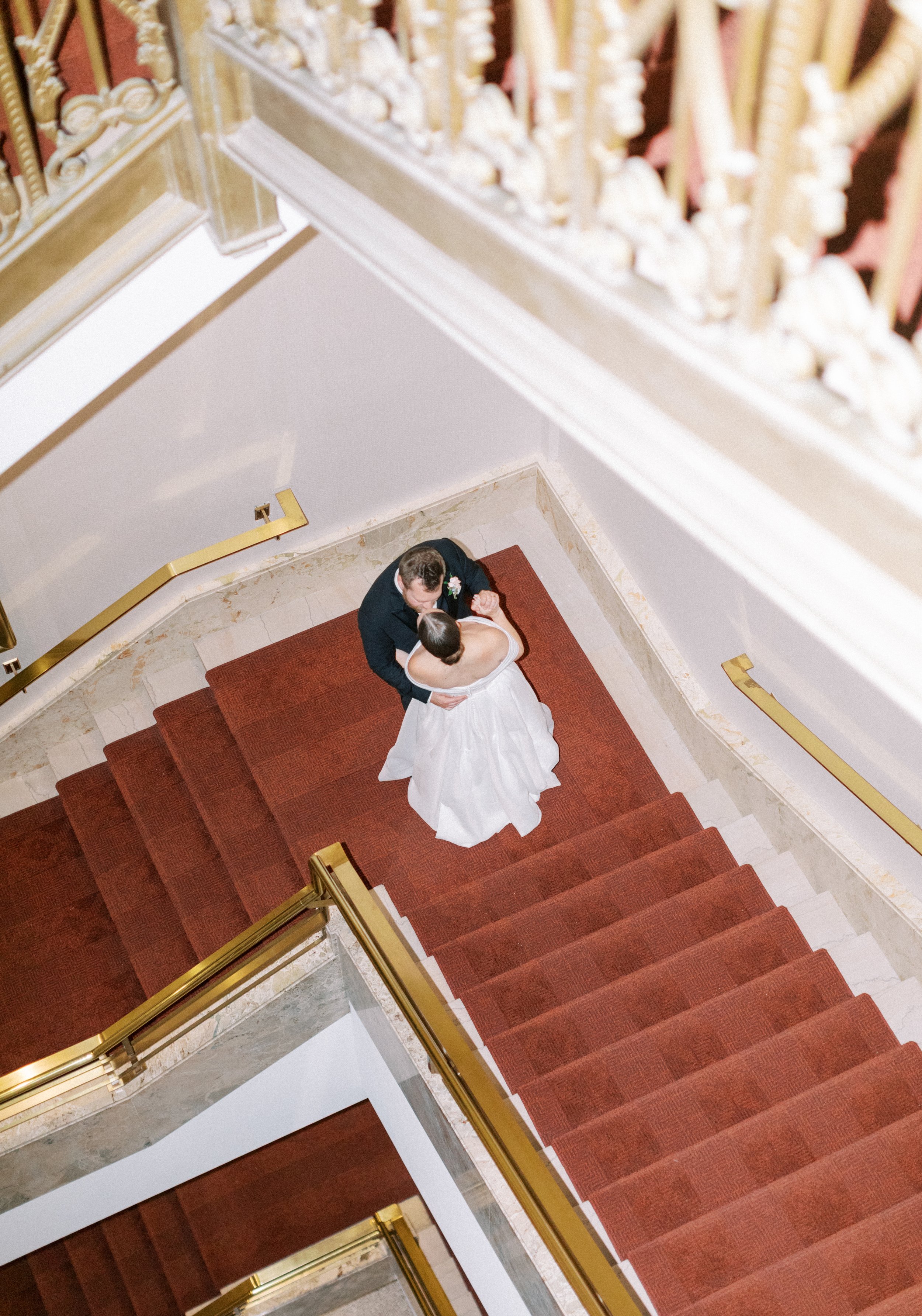 A bride and groom dancing on a staircase with ornate white and gold railing. The bride is wearing a white wedding dress, and the groom is in a black suit. The staircase has red carpeted steps.