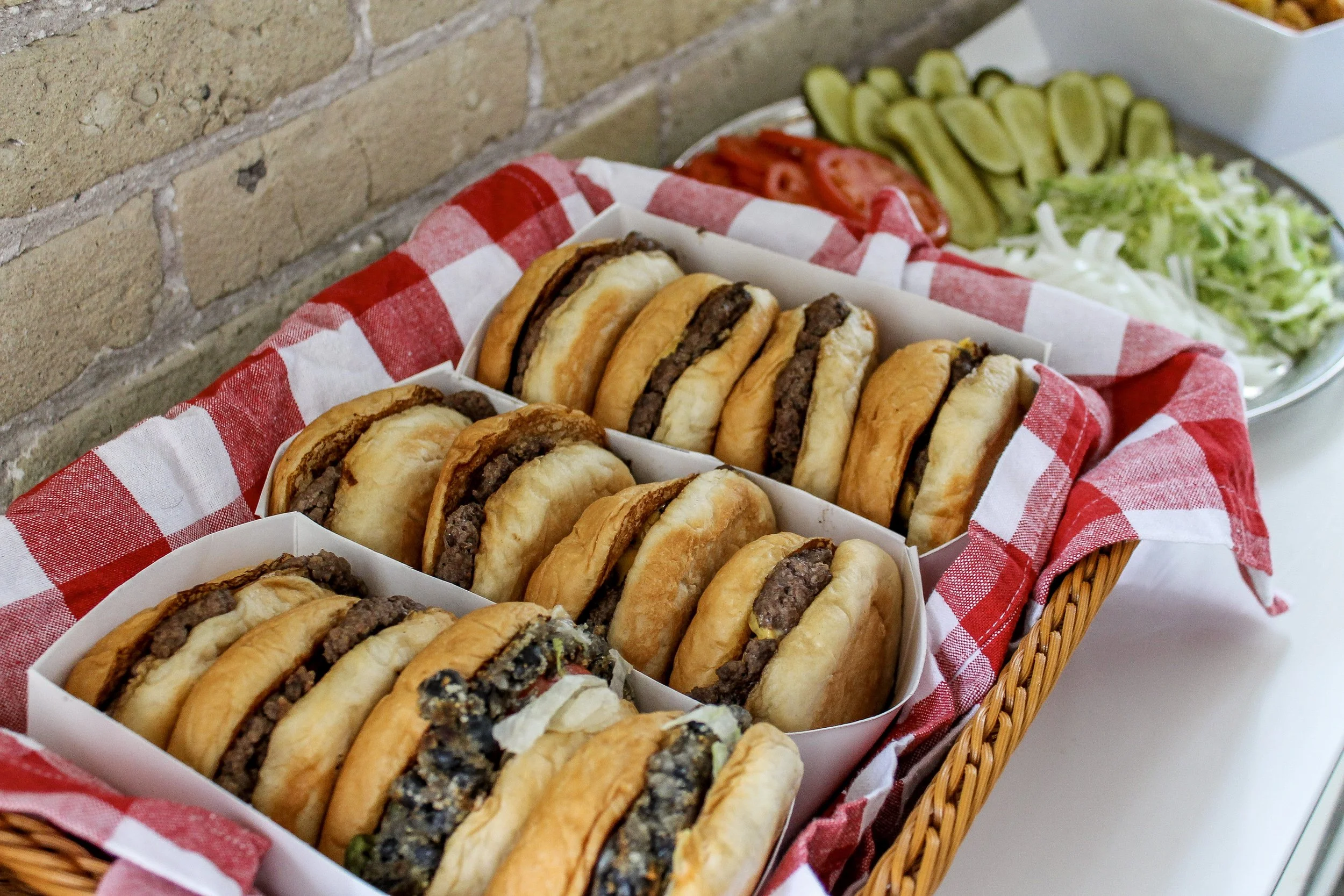 Basket of hamburgers on a red and white checkered cloth, with sliced pickles, tomatoes, onions, and lettuce in the background.