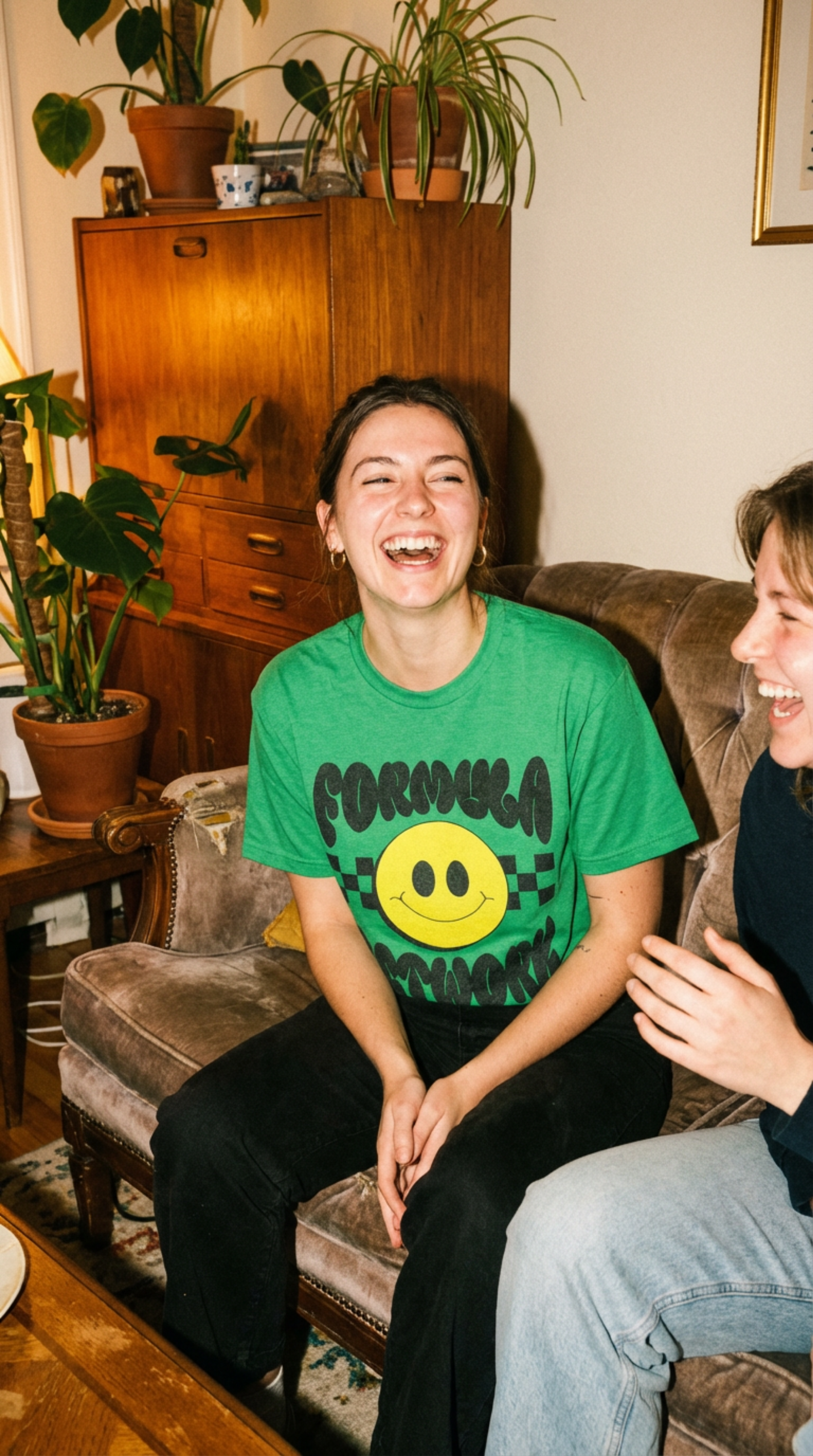 Two young women sitting on a vintage sofa, laughing and enjoying each other's company in a cozy living room with potted plants and wooden furniture.
