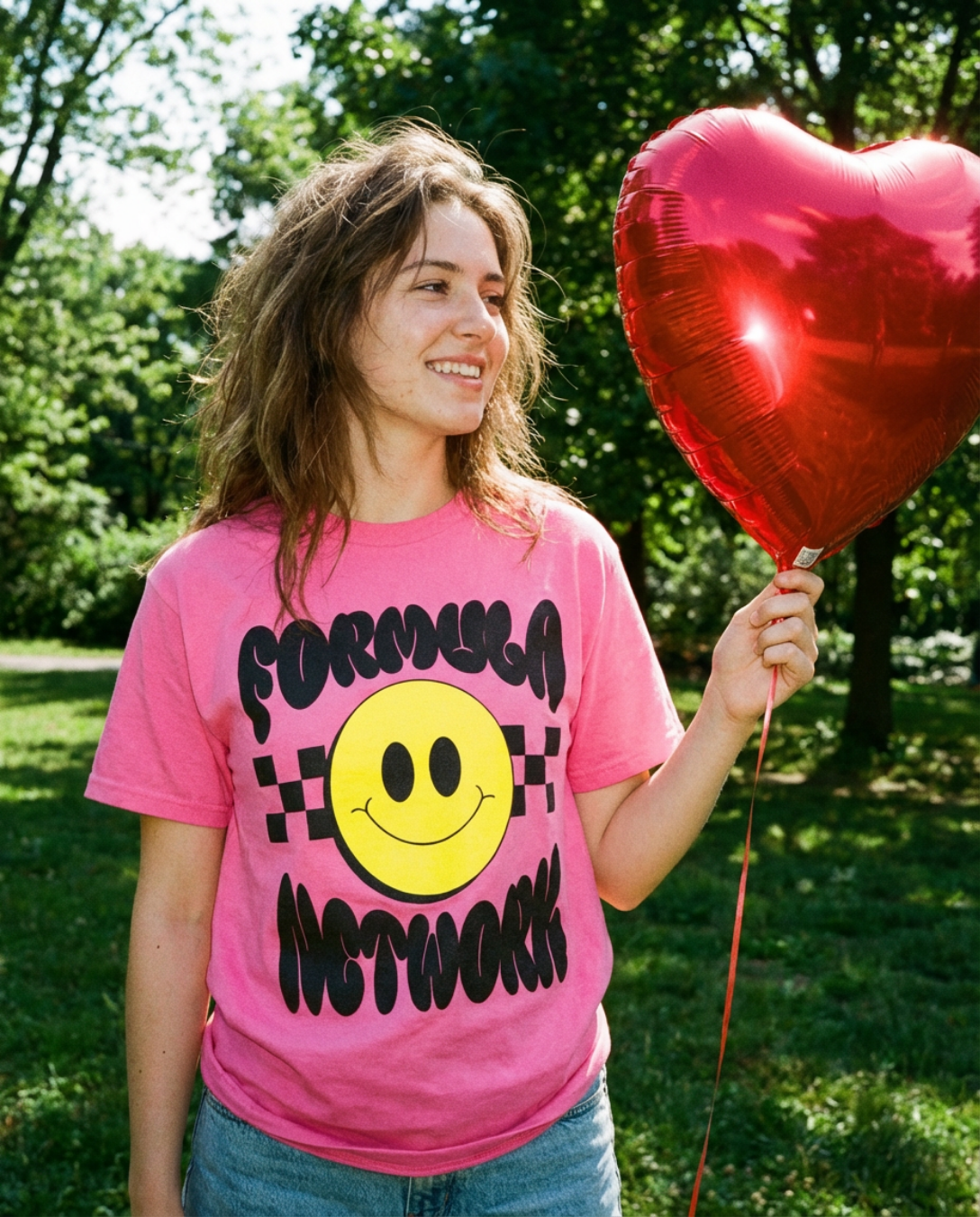 A young woman outdoors holding a red heart-shaped balloon, wearing a pink Formula Network T-shirt with a smiley face and text, smiling in a park with green trees and grass.