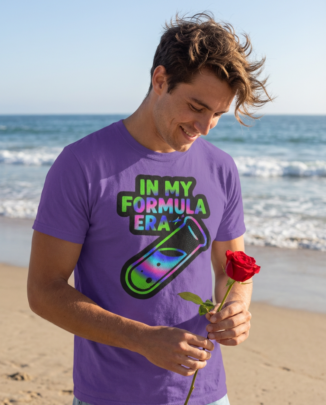 A smiling man in a Formula Network purple t-shirt with colorful text and graphic, holding a red rose on the beach with the ocean in the background.