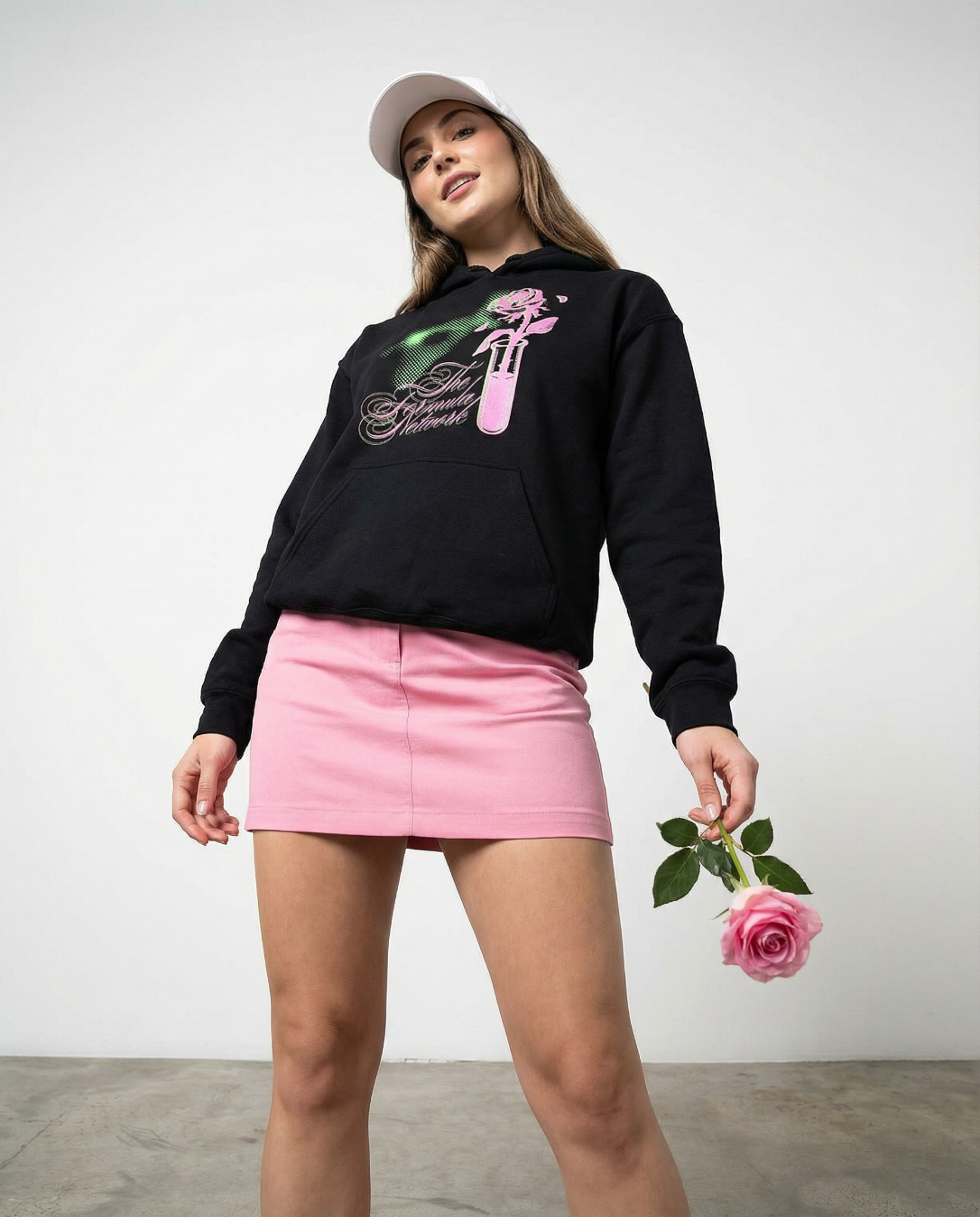 Young woman wearing a Formula Network black hoodie with a pink and green graphic, pink skirt, and white cap, holding a pink rose against a plain white background.