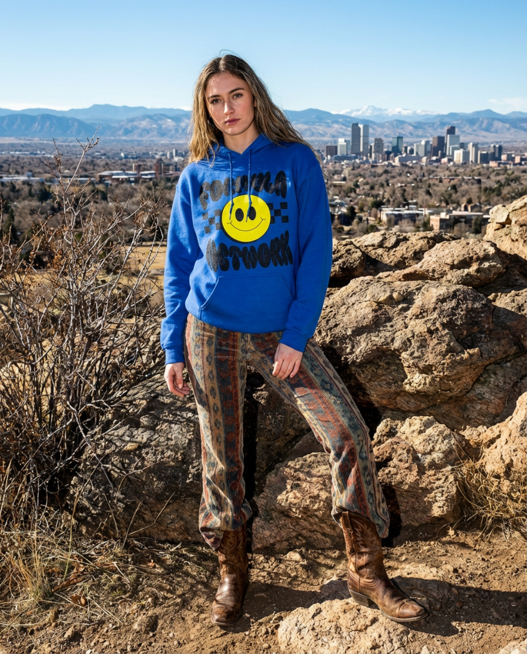 Young woman standing outdoors on rocks, wearing a bright blue hoodie with a Formula Network yellow smiley face graphic and text, patterned pants, and cowboy boots, with a cityscape and mountains in the background.