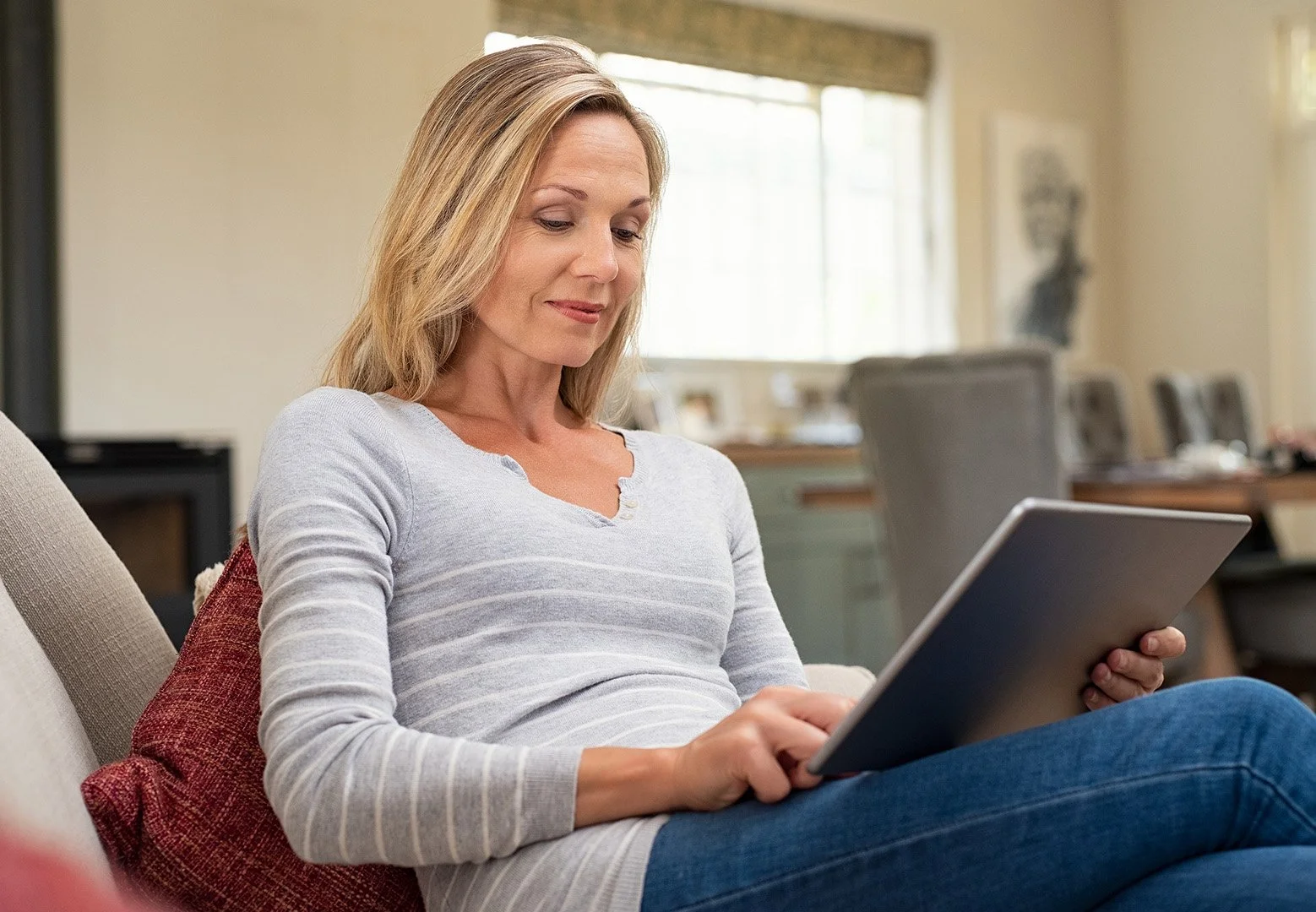 A woman with blonde hair sitting on a couch, looking at a tablet in her hands, in a well-lit home interior.