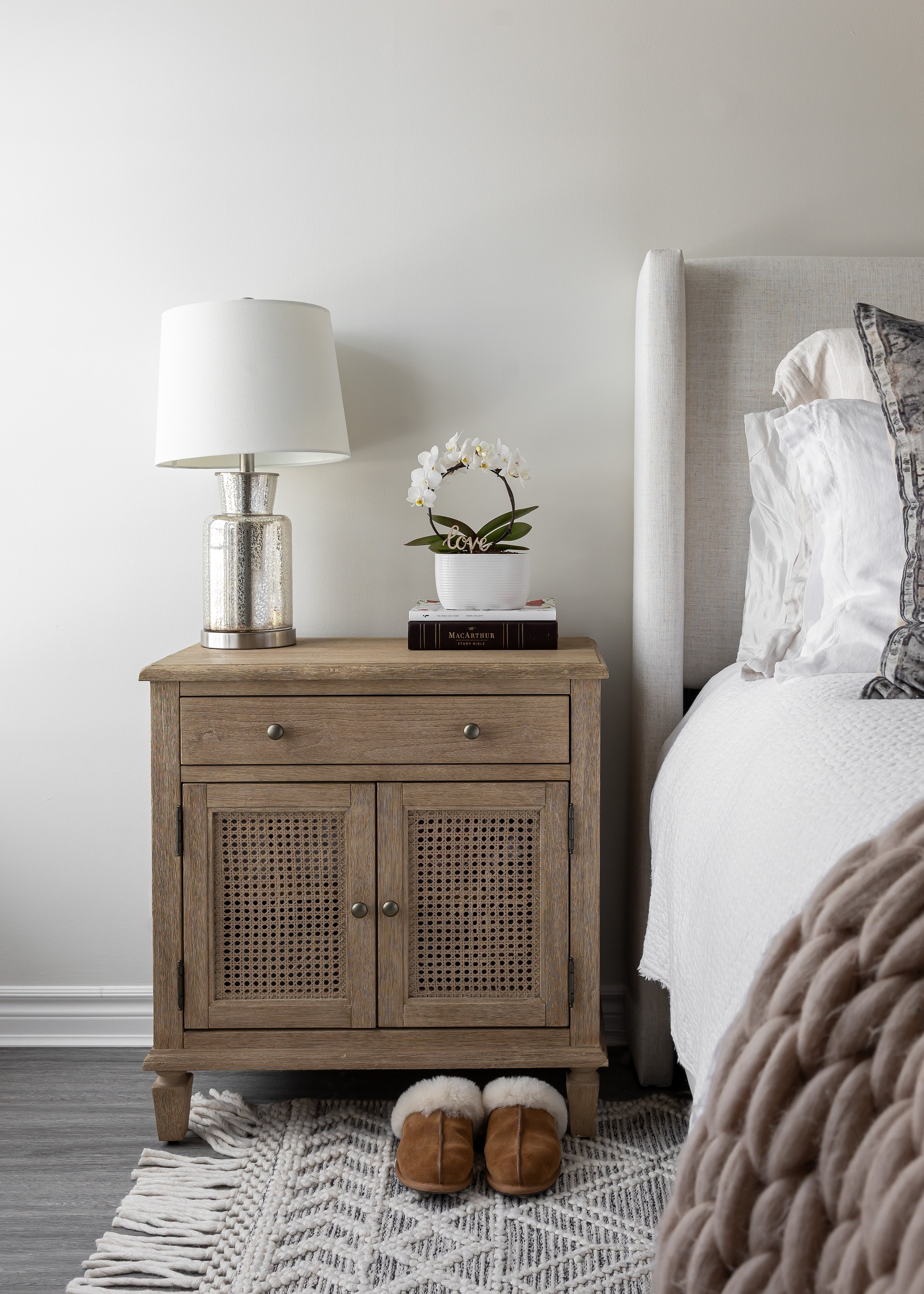 Modern farmhouse bedroom with a rustic wood nightstand featuring woven cane cabinet doors, a silver glass table lamp with a white shade, white orchid in a ceramic pot, stacked hardcover books, and shearling-lined slippers on a neutral geometric tasse