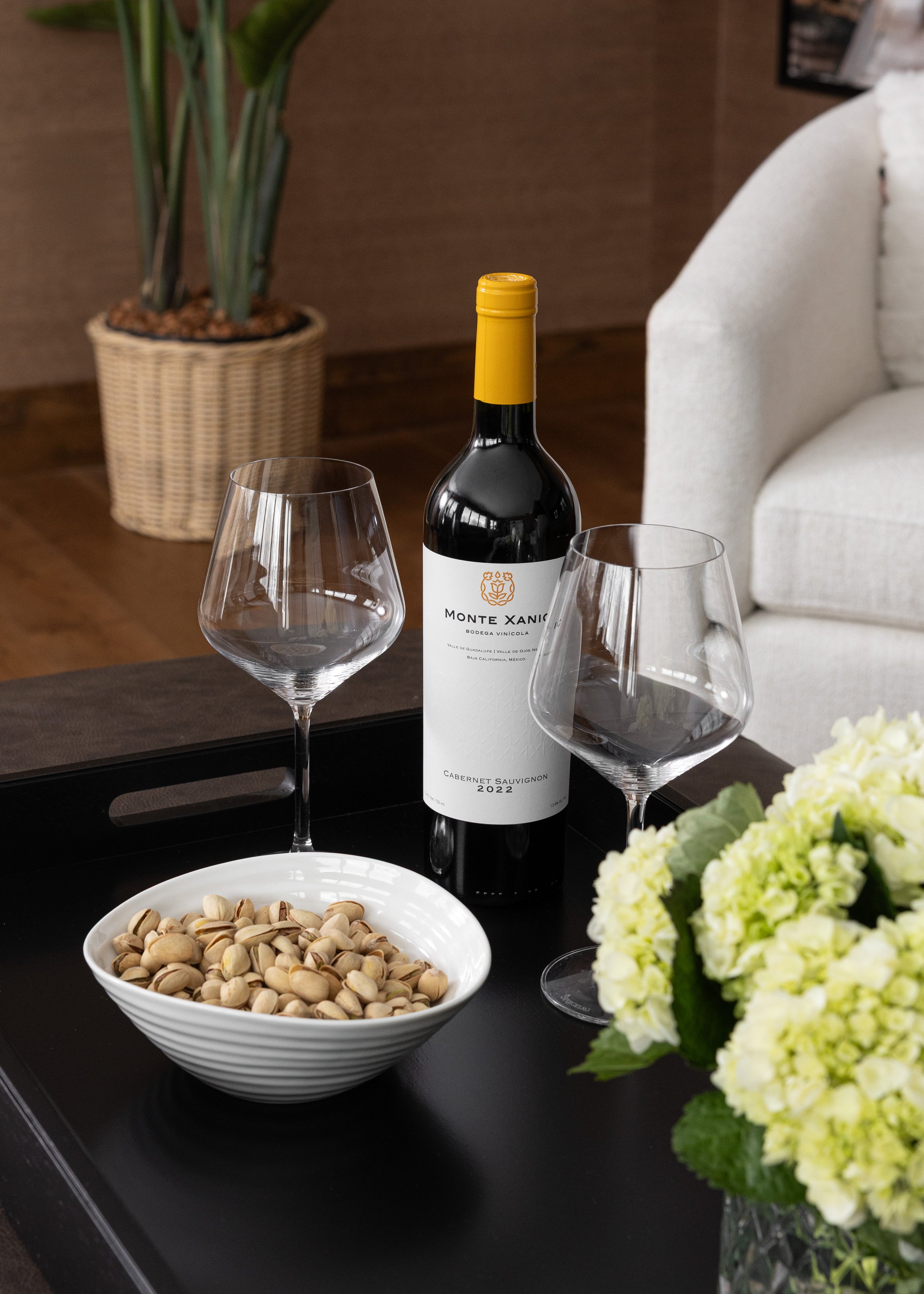Close-up of a cozy coffee table vignette featuring a bottle of Monte Xanic Cabernet Sauvignon 2022 with two elegant wine glasses, a white bowl of pistachios, and a fresh arrangement of white hydrangeas. The black tray contrasts beautifully against a