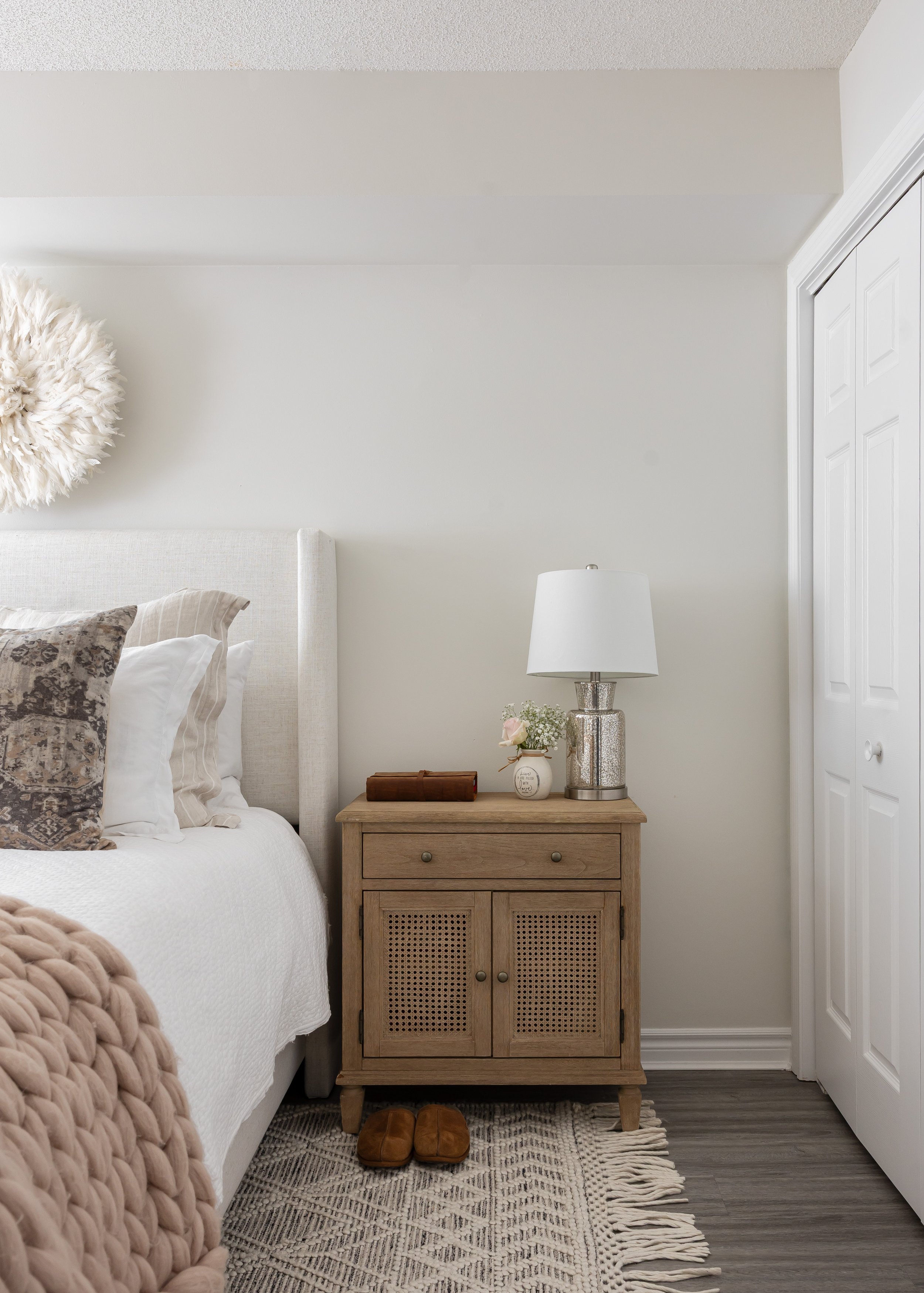 Modern farmhouse bedroom corner styled by Studio 7 Designs featuring a light oak nightstand with woven cane doors, a silver mercury glass lamp with white shade, fresh flowers in a bud vase, and cozy decor touches like a knit throw, plush slippers, an