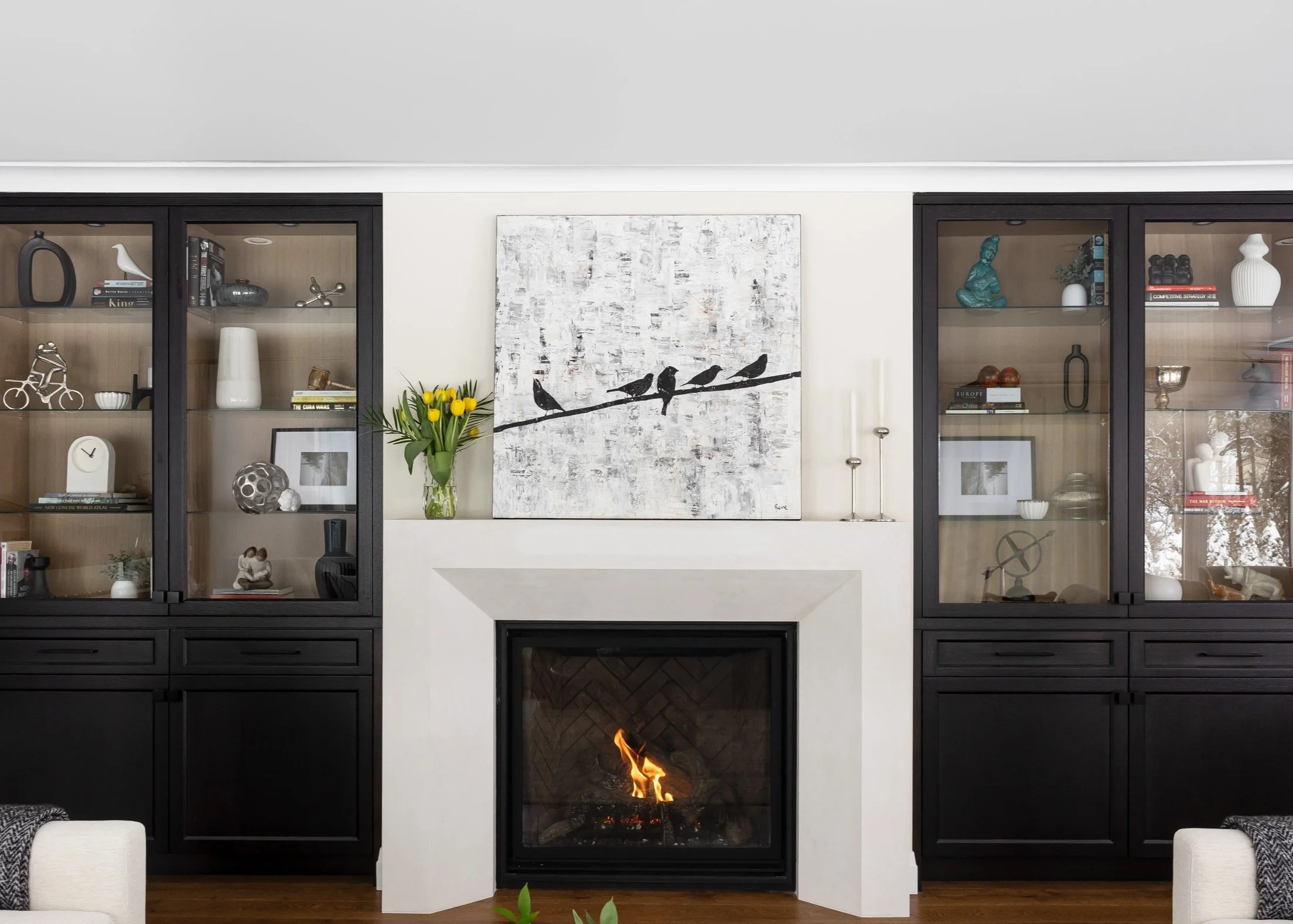 A living room with two black cabinets with glass doors on either side of a white fireplace. On the wall above the fireplace is a white and black painting of birds sitting on a branch. The cabinets display decorative objects and books. On the mantle is a vase with yellow tulips.
