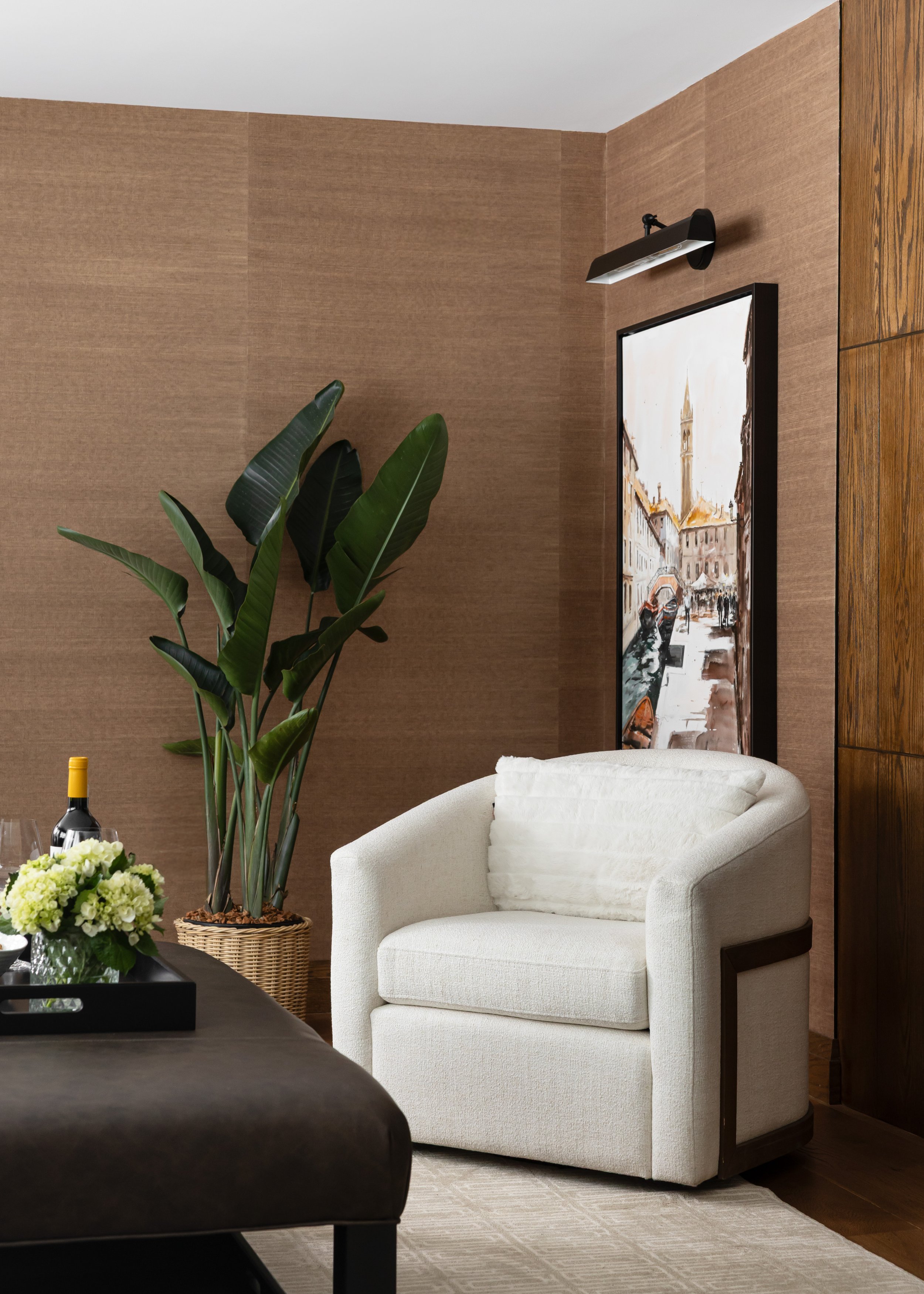 Cozy reading nook designed by Studio 7 Designs featuring a modern white barrel chair with wood accents, moody gallery-style wall lighting above European-inspired artwork, and natural textures layered with soft neutrals for a timeless, serene aestheti
