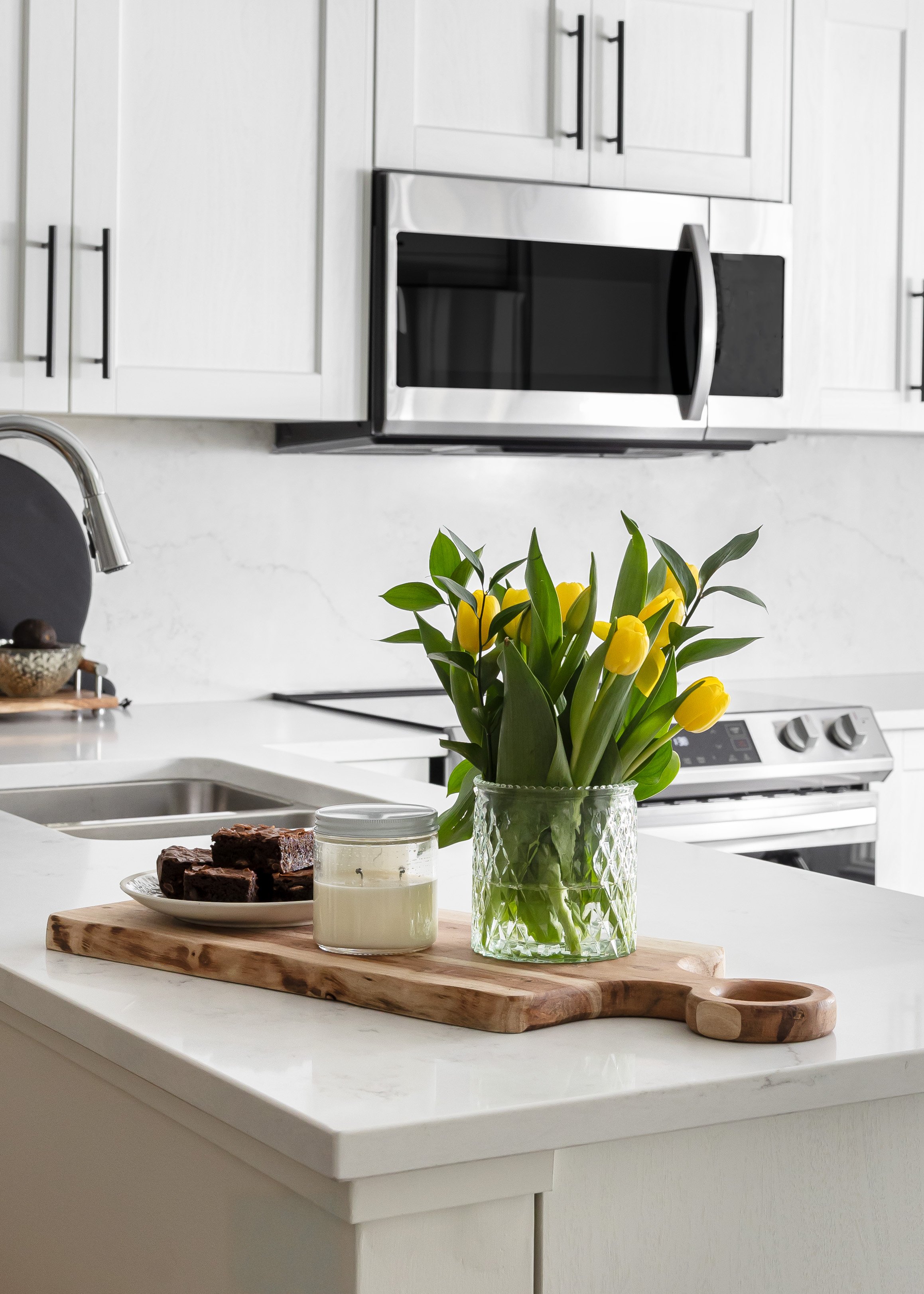 Bright modern kitchen featuring a white quartz waterfall island styled with a wooden cutting board, fresh yellow tulips in a textured glass vase, a white soy candle, and a plate of chocolate brownies. Studio 7 Designs decor blends minimalism with war