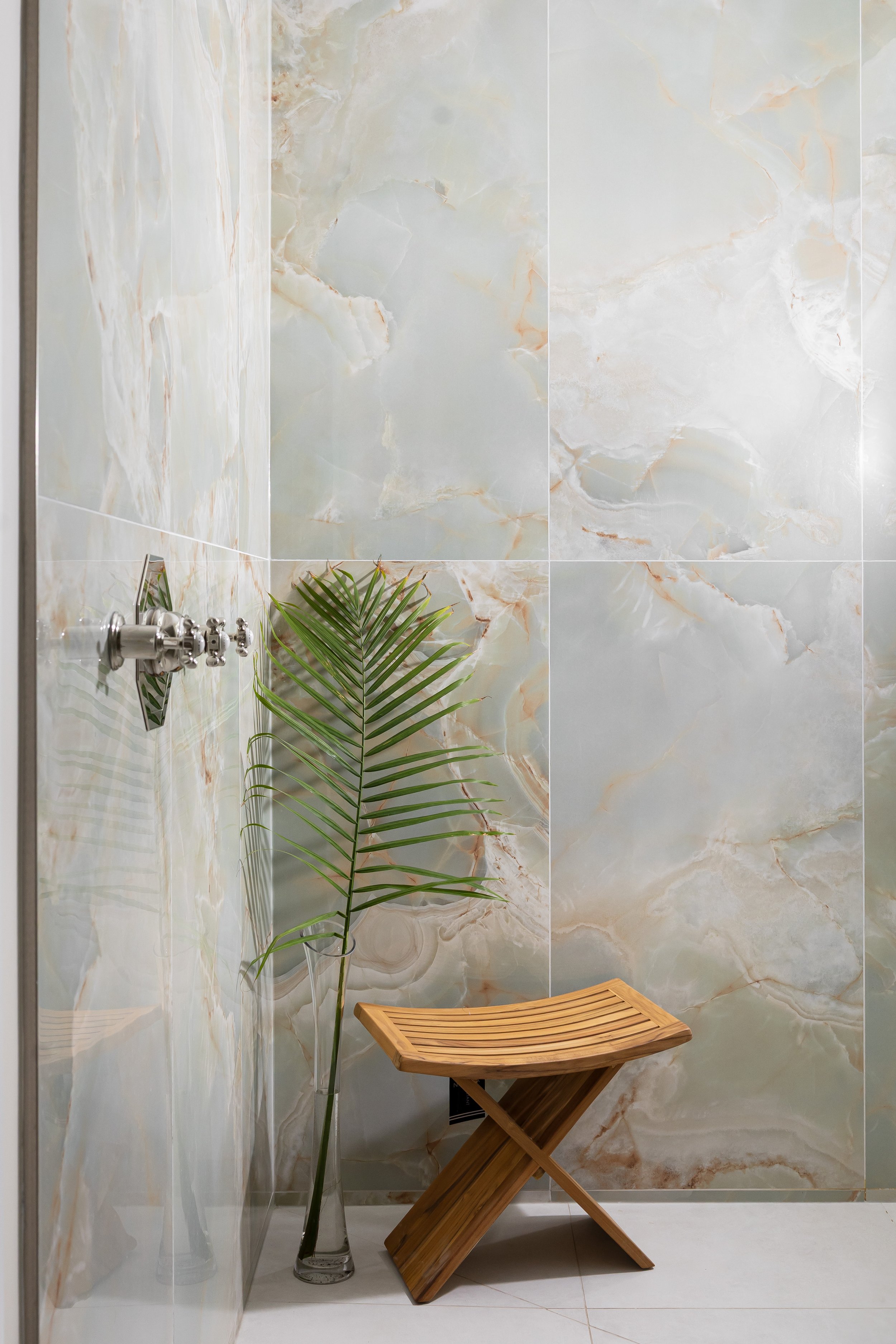Spa-inspired shower design featuring large format onyx-look tiles with soft green and amber veining. A minimalist wooden stool and a tall glass vase with a single palm leaf create a serene, organic touch. Designed by Studio 7 Designs to evoke a luxur