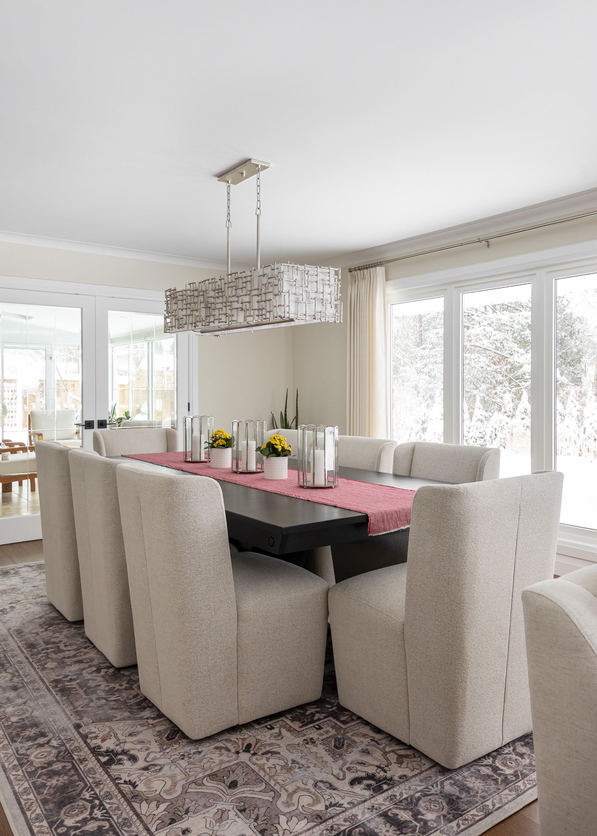 Elegant dining room by Studio 7 Designs featuring a dark rectangular dining table surrounded by modern cream upholstered chairs. A soft red table runner with glass candle holders and yellow potted flowers adds a soft touch, complemented by a geometri