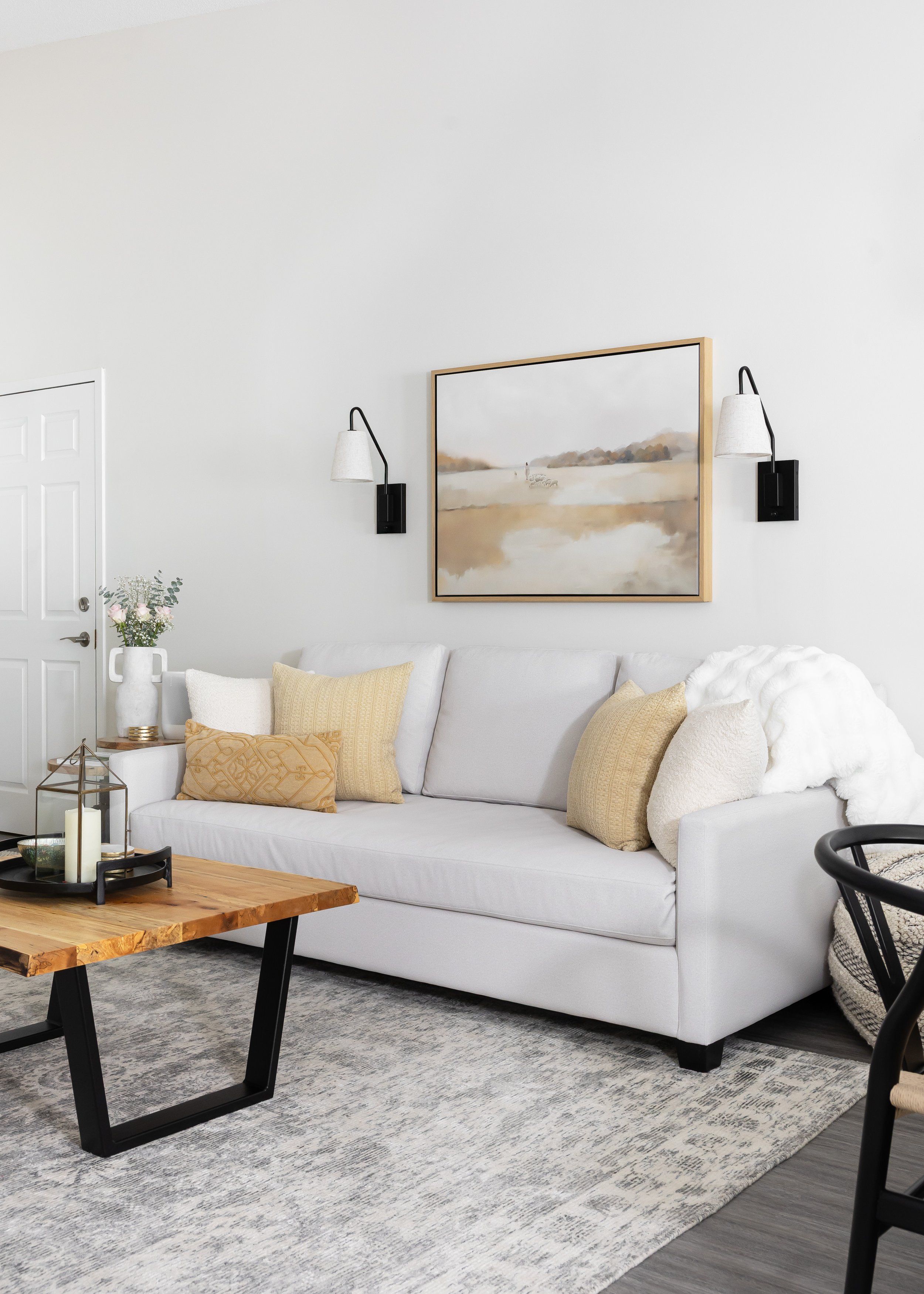 Neutral modern living room designed by Studio 7 Designs featuring a light gray sofa with textured throw pillows, a rustic live-edge wood coffee table with black metal legs, a layered area rug, soft white throw blanket, and landscape artwork framed be
