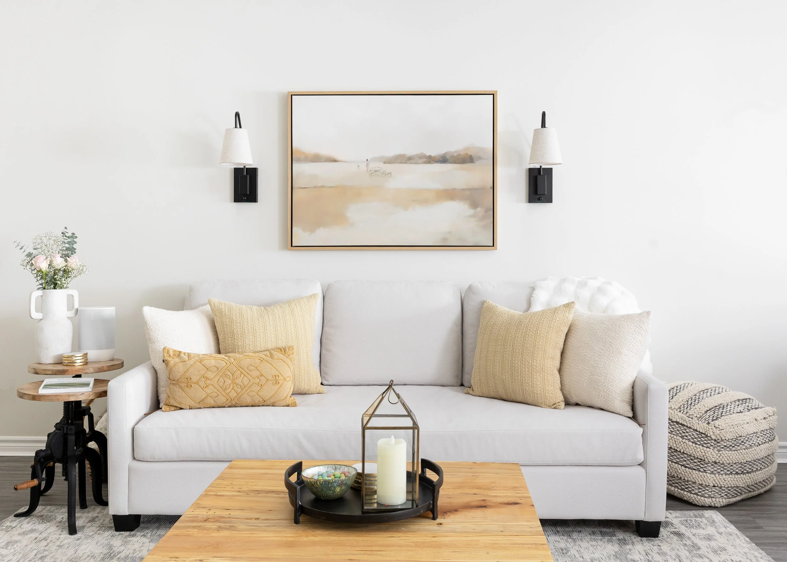 Studio 7 Designs Custom Home of Living room with a white sofa, beige pillows, a wooden coffee table with candles and decorative items, a wall art painting, two wall-mounted lights, a side table with a flower vase, and a pile of blankets to the right.