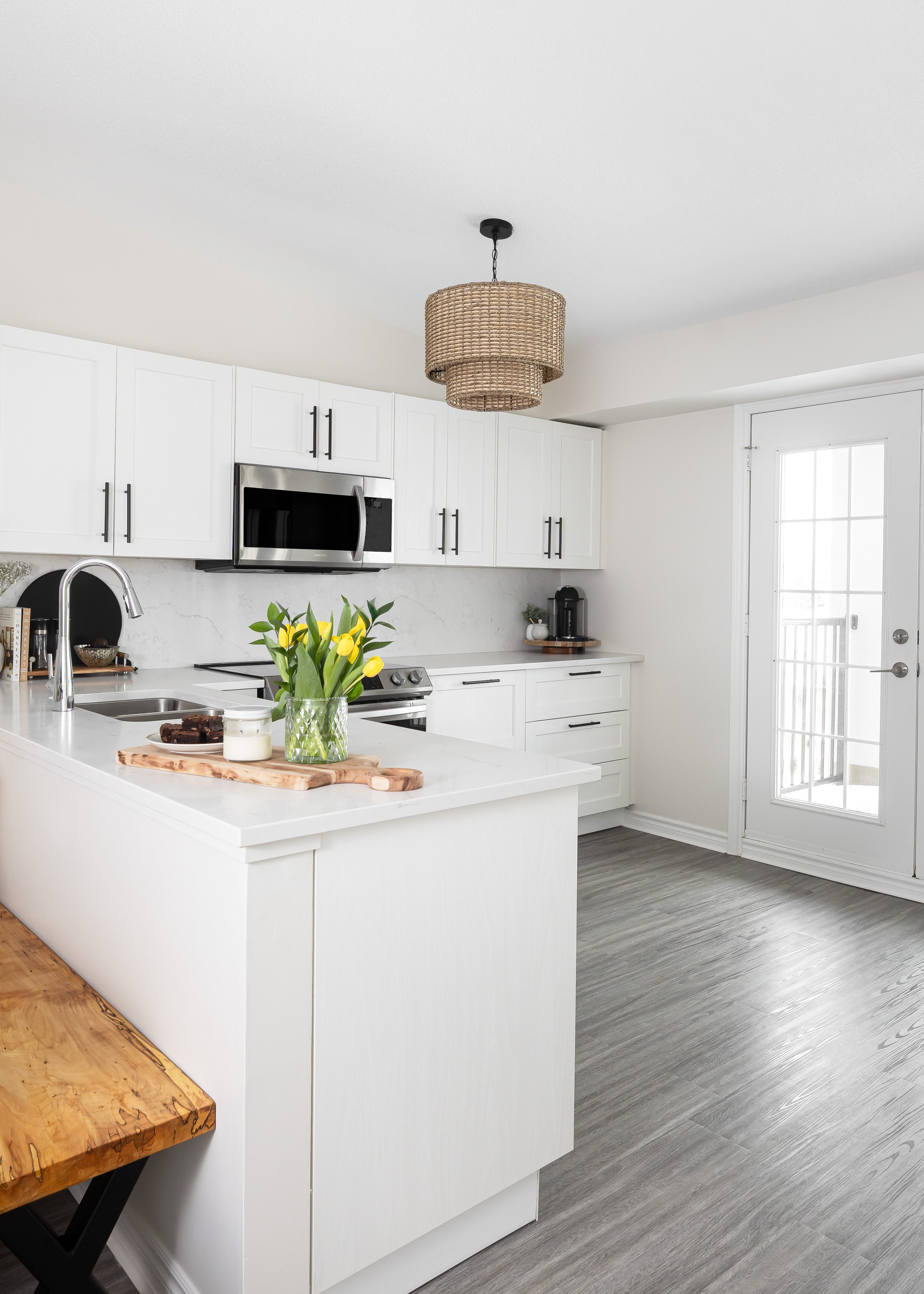 Bright and modern white kitchen designed by Studio 7 Designs, featuring white shaker cabinets with black handles, quartz countertops, stainless steel appliances, a rattan pendant light, and a kitchen island styled with fresh yellow tulips and natural