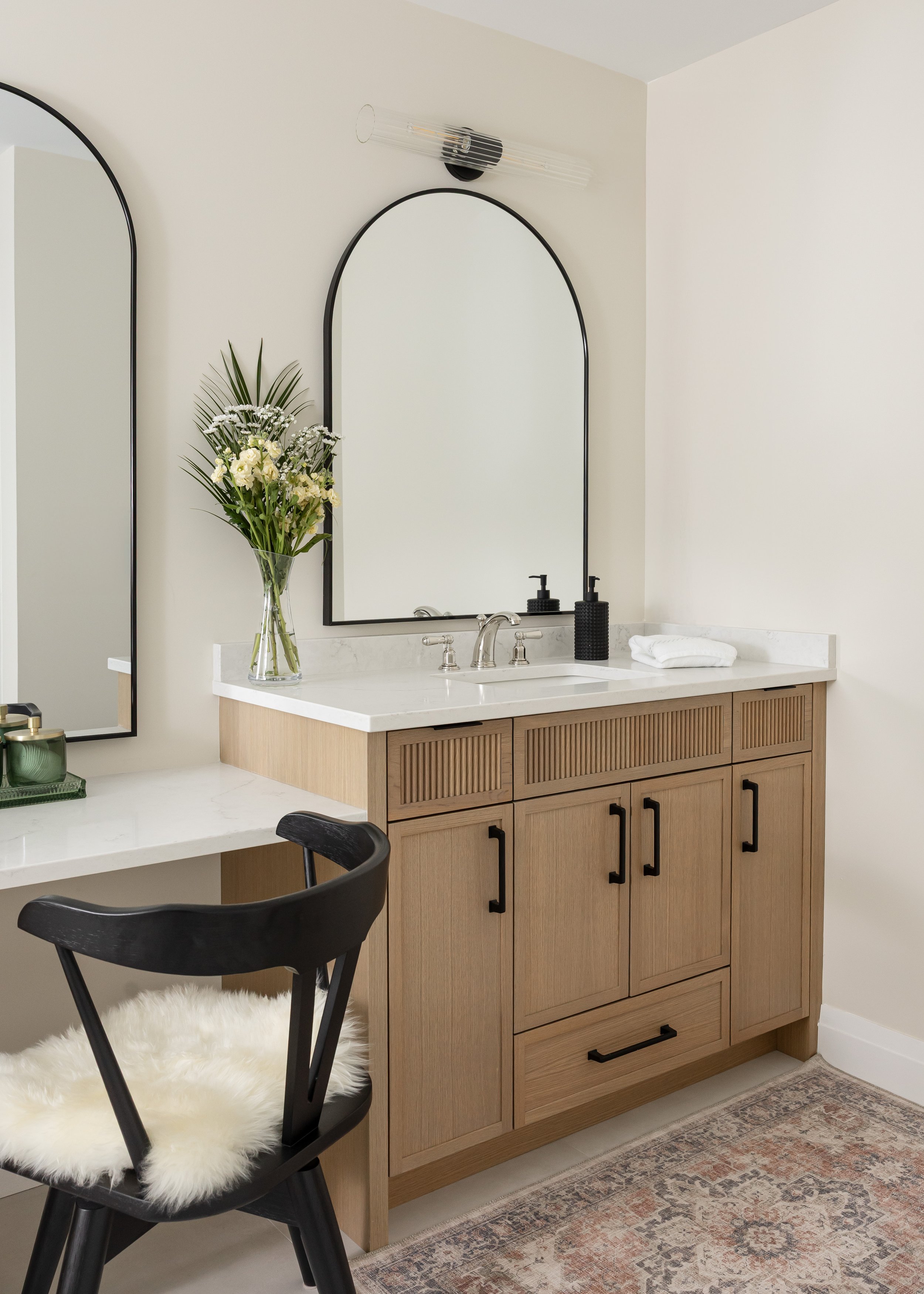 Modern wood vanity with white quartz countertop and black hardware, paired with an arched black-framed mirror and wall-mounted glass sconce. A cozy makeup seat with a black spindle-back chair and faux fur cushion adds charm, while fresh flowers in a