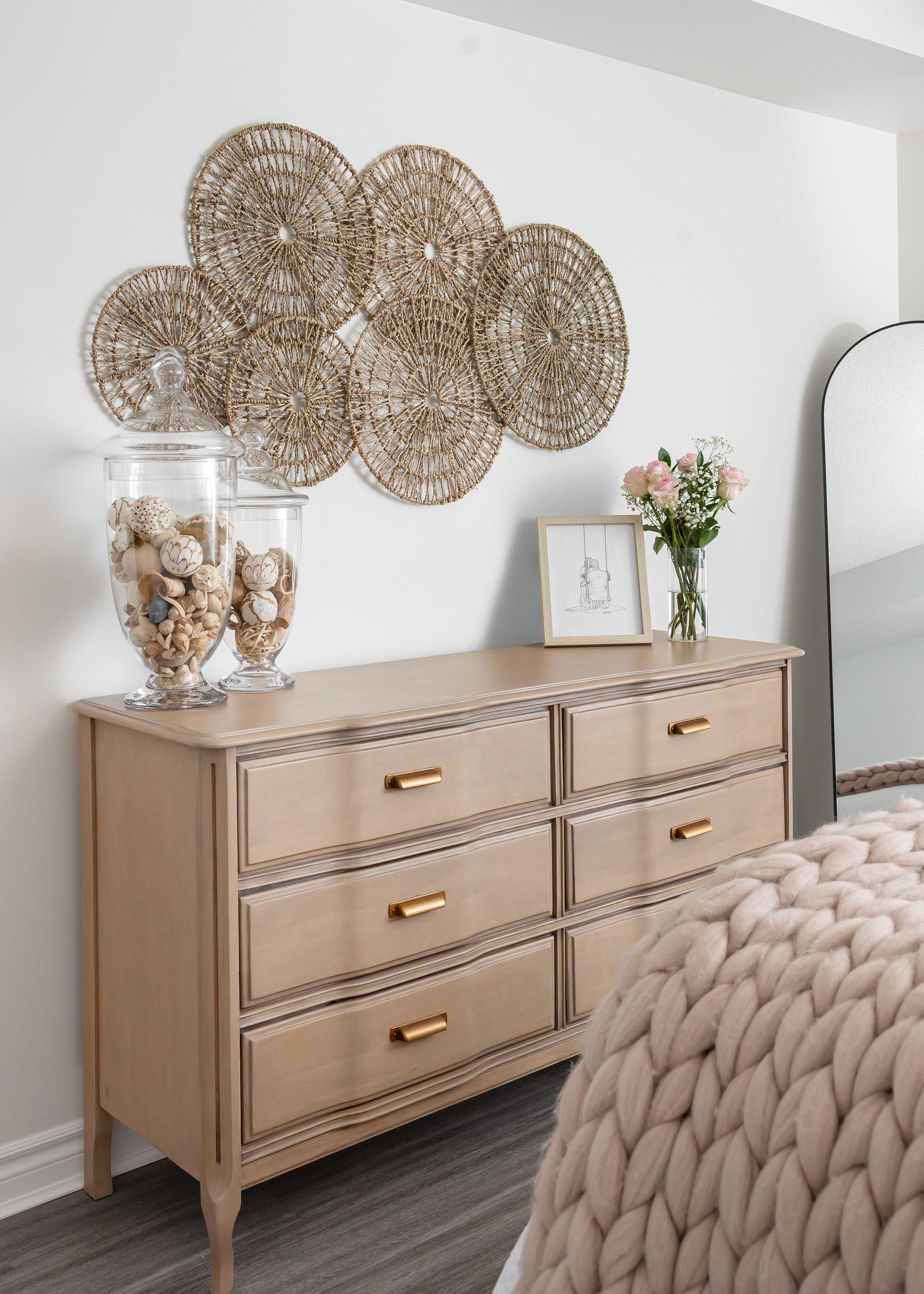 Neutral bedroom with a light oak 9-drawer dresser featuring brass handles, styled with coastal-inspired apothecary jars, pink roses in a glass vase, and a modern line art frame. A boho woven rattan wall decor adds texture above, styled by Studio 7 De