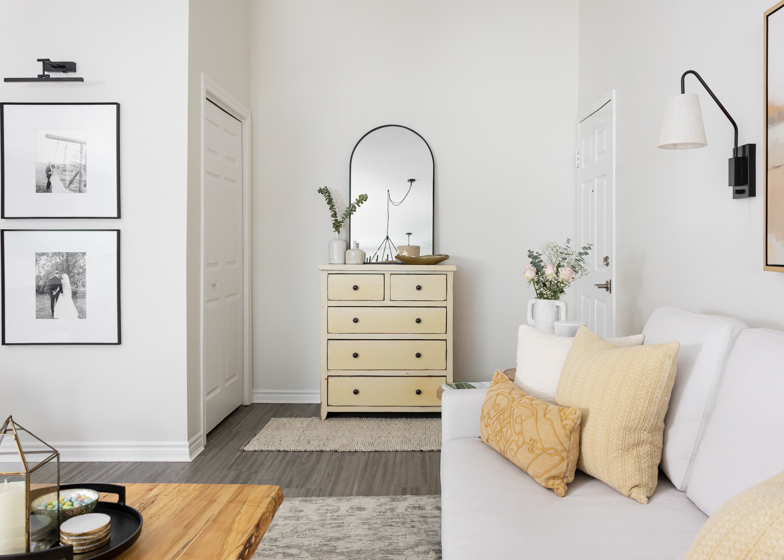Modern living room corner styled by Studio 7 Designs, featuring a soft white sofa with cream and mustard accent pillows, vintage-style ivory dresser with black knobs, arched black-framed mirror, decorative ceramics, and framed black-and-white photogr