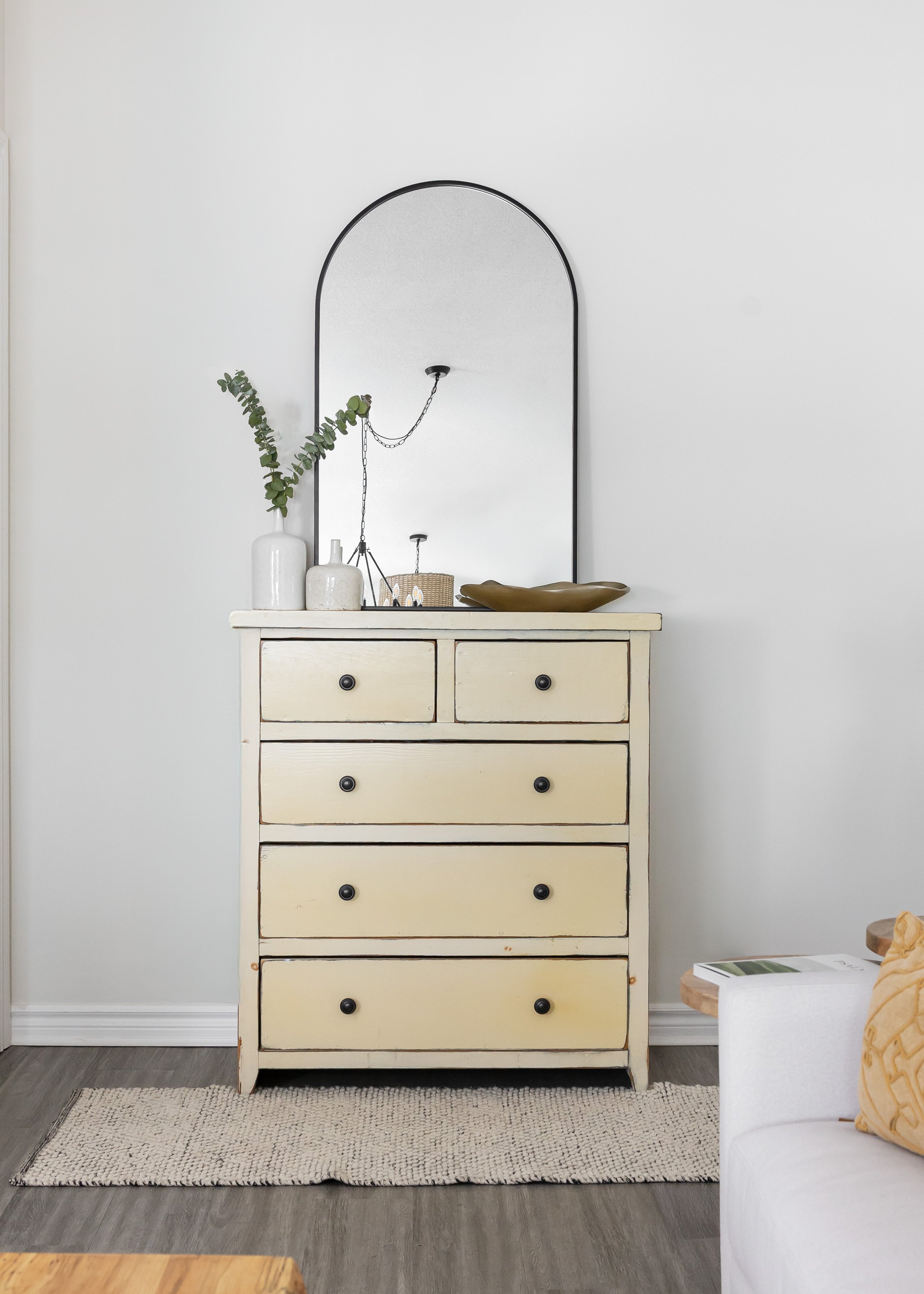 Styled neutral vignette featuring a vintage cream dresser with black hardware, topped with ceramic vases, a sculptural bowl, and eucalyptus stems, paired with an arched black-framed mirror reflecting modern lighting—Studio 7 Designs' refined blend of