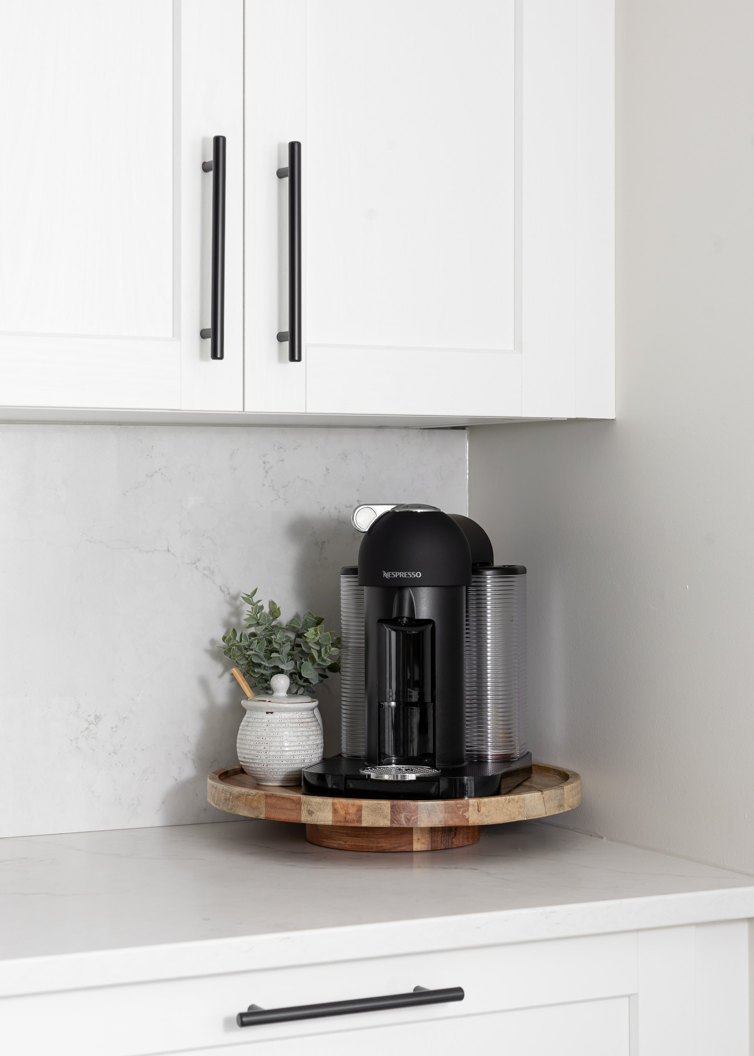 Modern kitchen coffee station with a sleek black Nespresso machine on a round wooden tray, paired with a ceramic jar holding stir sticks and faux greenery. Styled on white quartz countertops with shaker-style white cabinets and matte black hardware.