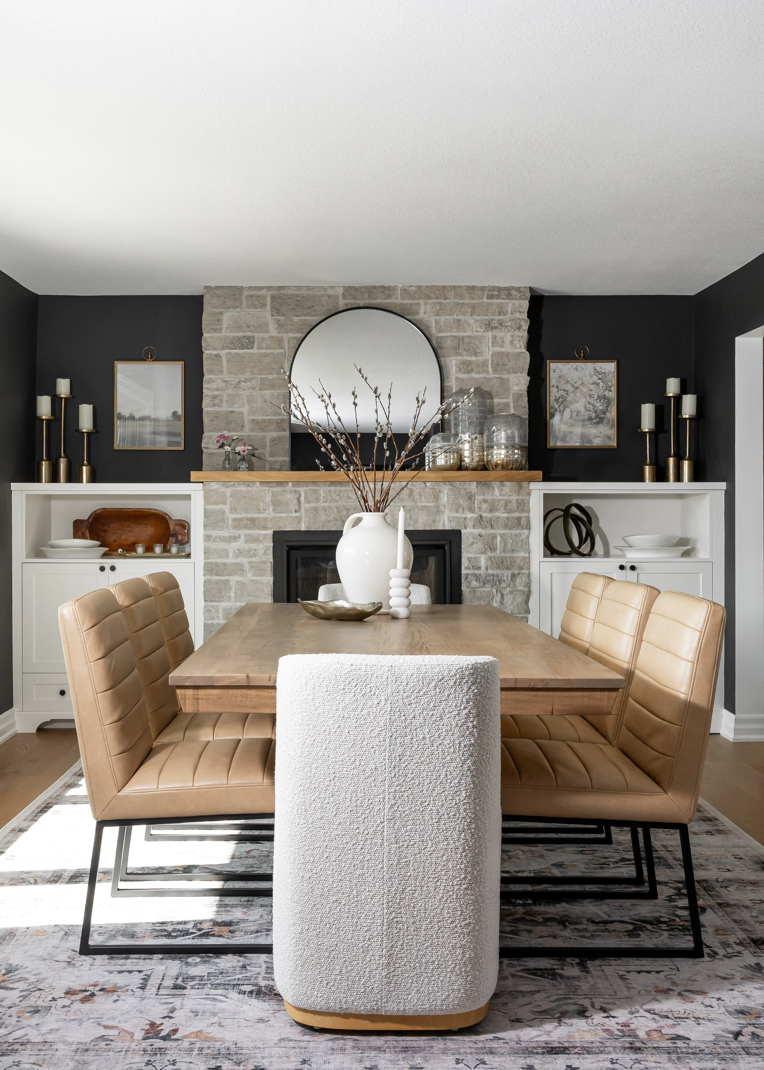 Studio 7 Designs Custom dining room with a wooden table, tan upholstered chairs, a white textured armchair, and a white and tan rug. The background features a stone fireplace, a round mirror, framed artwork, and decorative items on white shelves.
