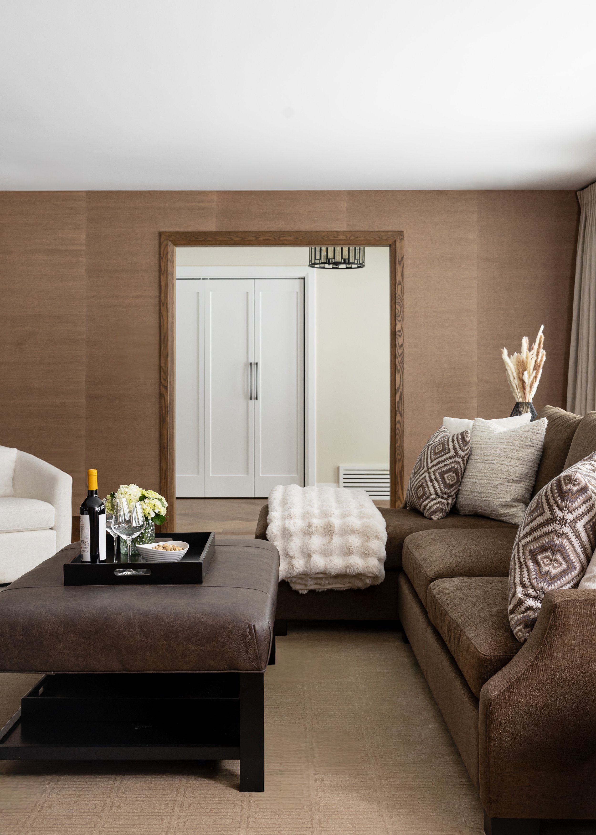 Sophisticated living room by Studio 7 Designs featuring textured neutral wallpaper, custom wood door trim, and a large leather ottoman styled with a black tray, wine, and florals—blending comfort with tailored elegance.
