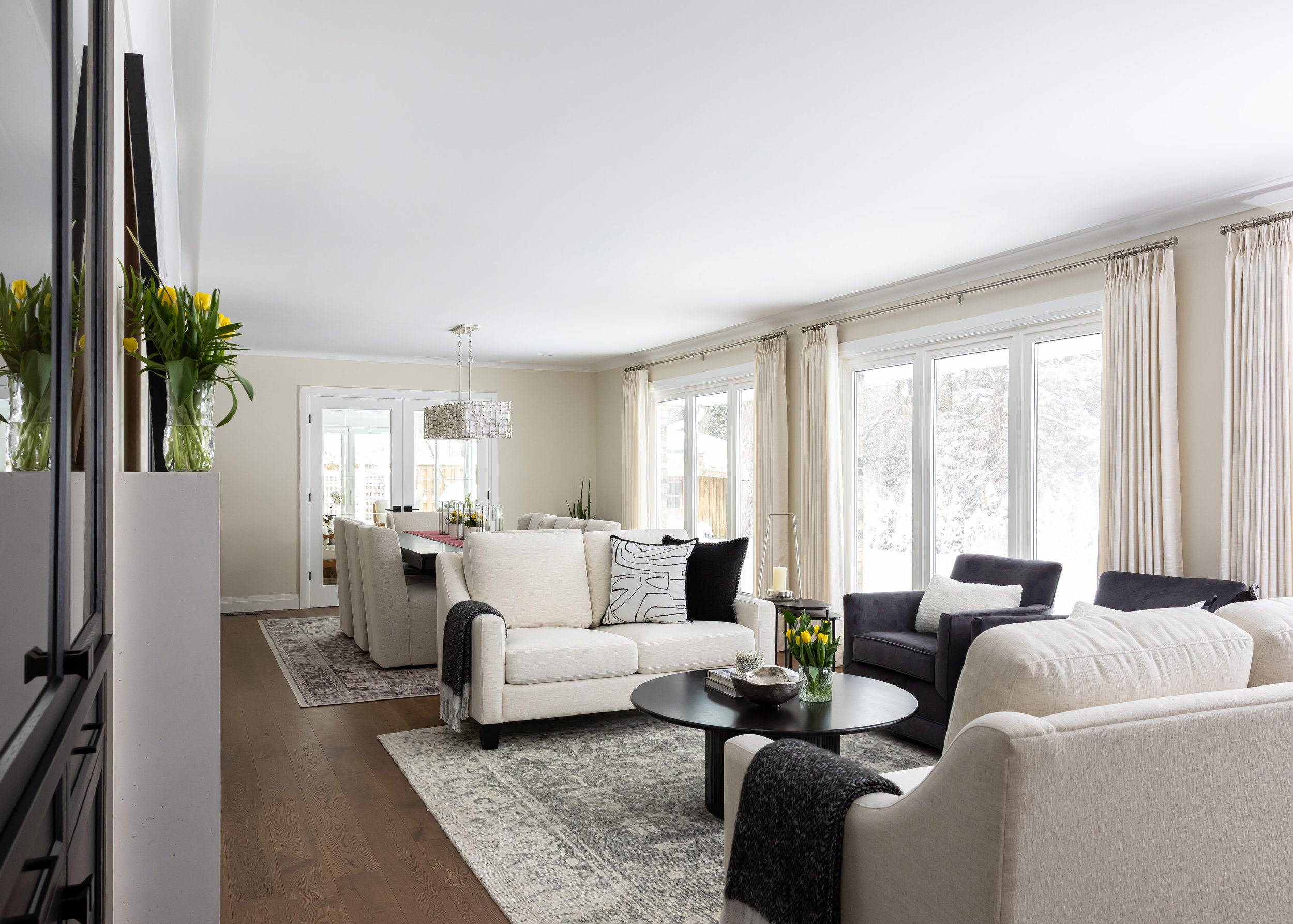 Bright and airy open concept living and dining room featuring cream armchairs, black accent chairs, and a round black coffee table layered over a vintage-style area rug. Floor-to-ceiling windows with soft ivory drapes flood the space with natural lig