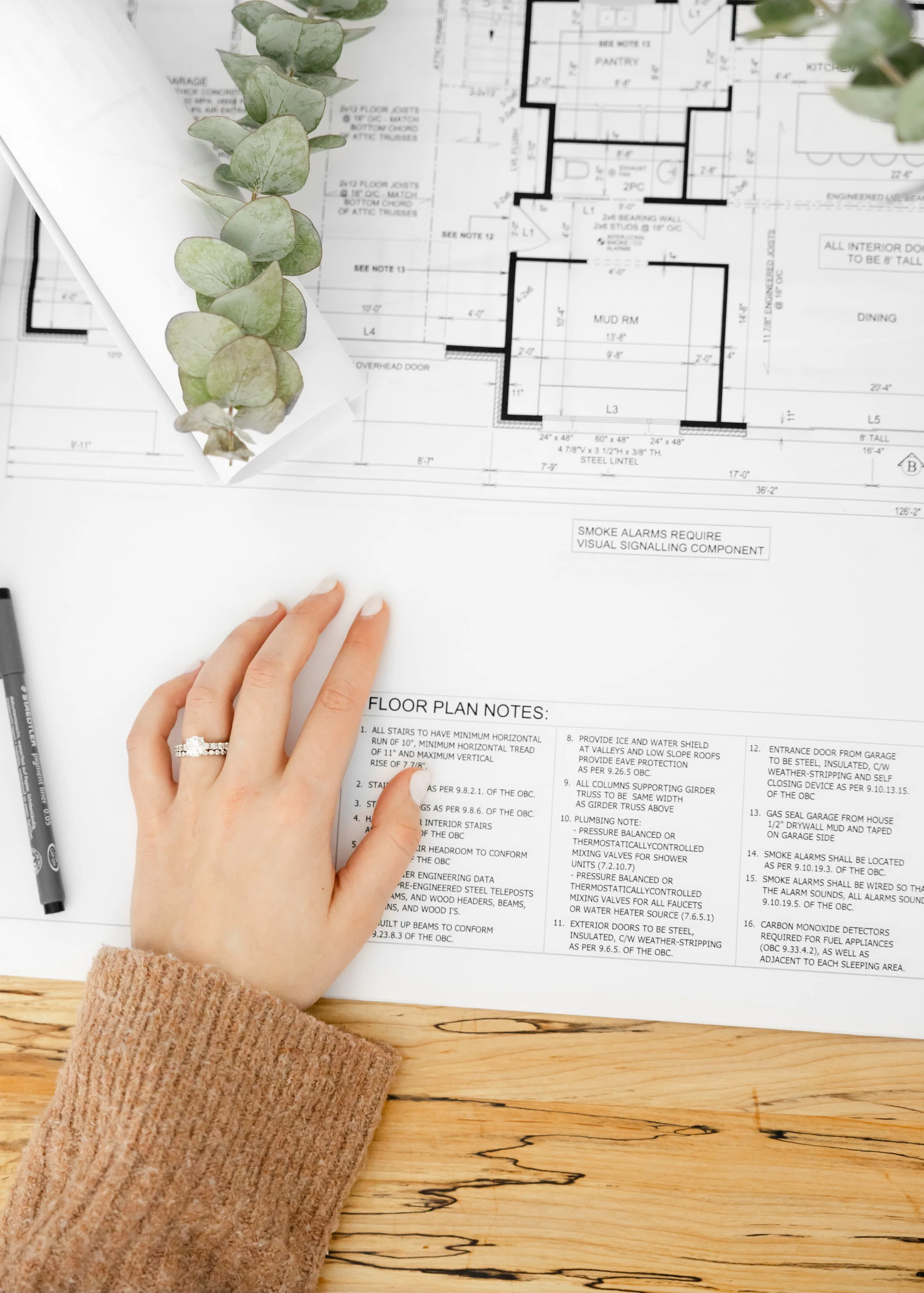 Close-up of a person’s hand with a ring, resting on a detailed architectural floor plan spread out on a wooden surface. The person is wearing a beige sweater, and there is a sprig of greenery on top of the floor plan. A black marker is visible near the bottom left corner of the image.