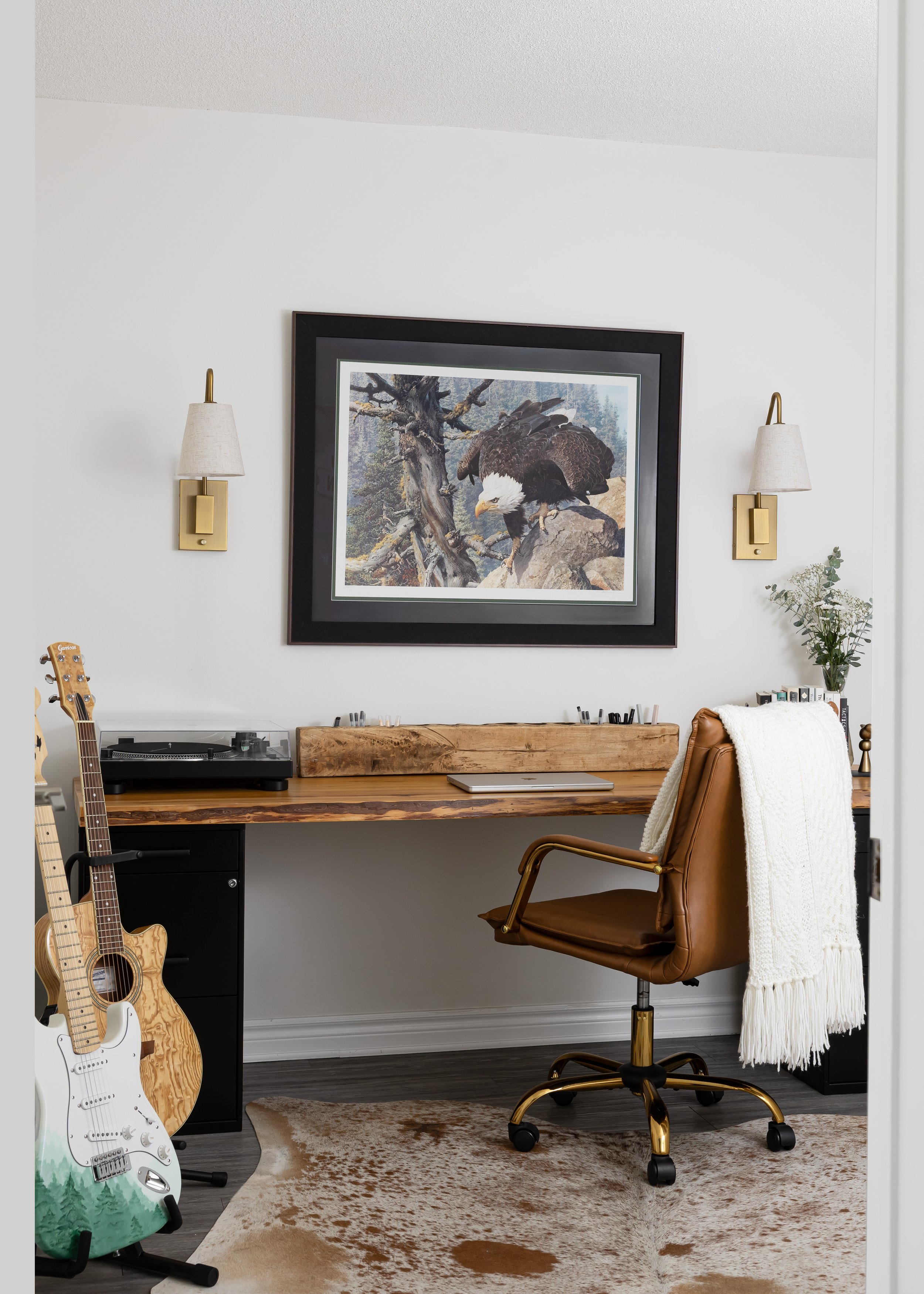 Rustic home office design with a live-edge wood desk, leather office chair, and nature-inspired decor—featuring a bald eagle painting and guitars for a creative vibe. Interior by Studio 7 Designs.