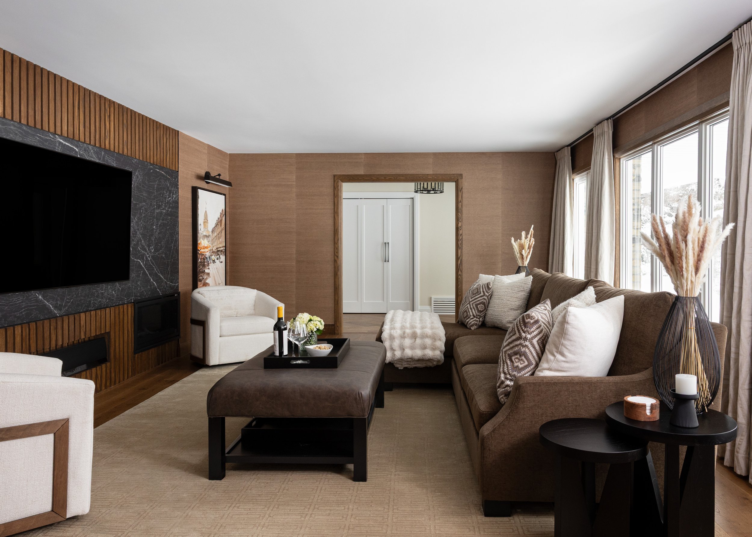 Elegant living room interior by Studio 7 Designs showcasing a cohesive design with a brown sectional sofa, textural throw pillows, large windows with blackout drapery, and a custom entertainment wall with a black marble fireplace.
