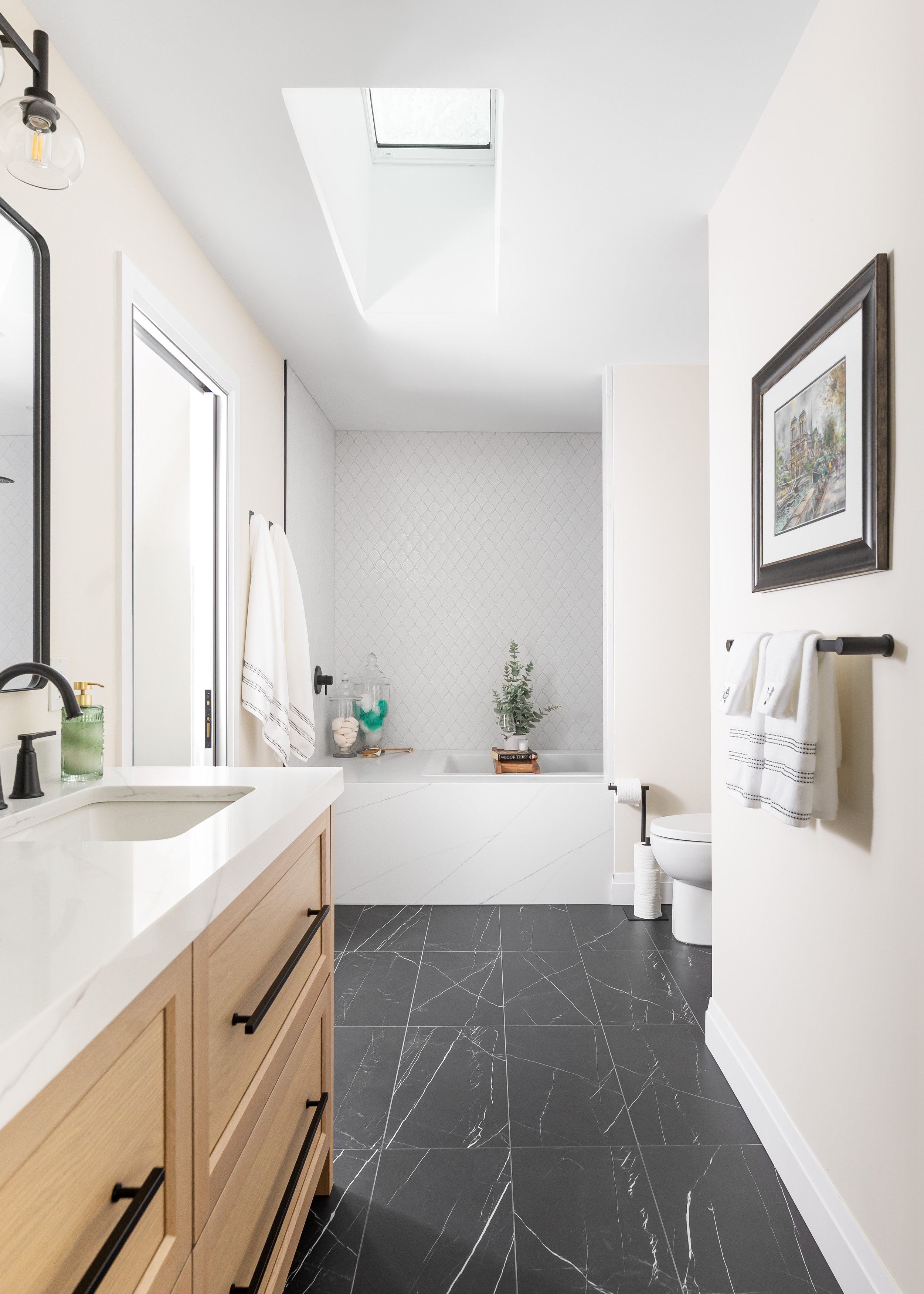 Modern full bathroom renovation by Studio 7 Designs featuring black marble tile flooring, natural wood double vanity, sleek black fixtures, and a soaking tub with textured white tile surround—chic and functional bathroom design inspiration.