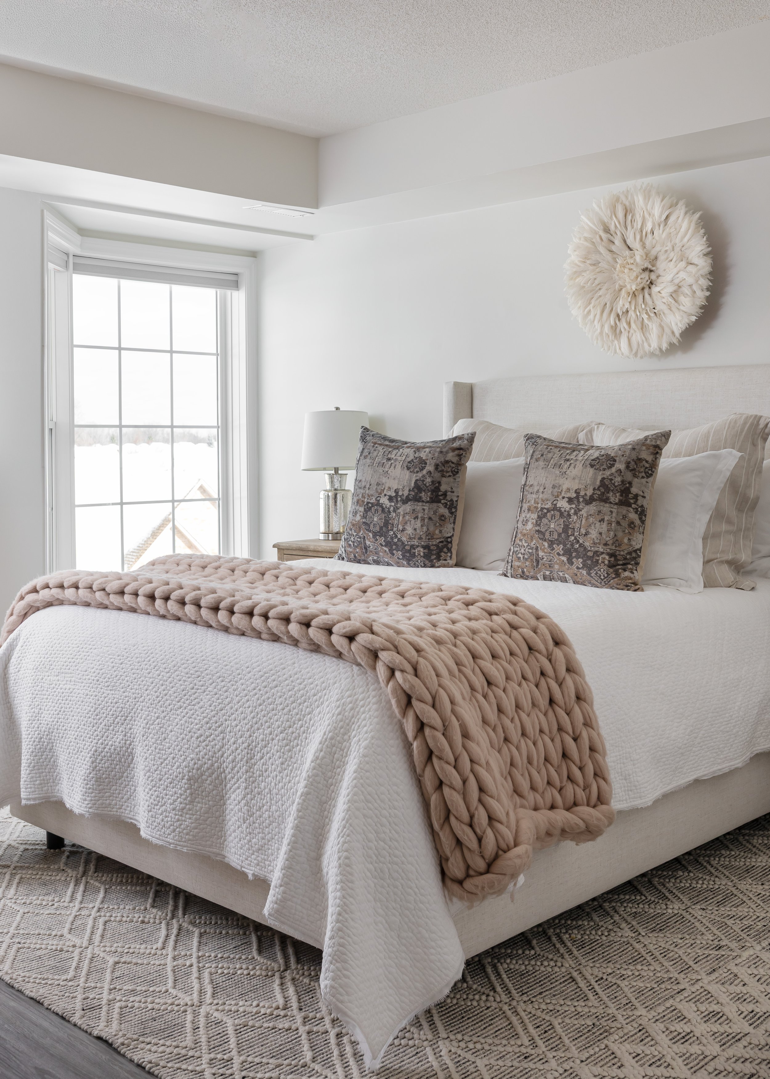 Bright and cozy bedroom styled by Studio 7 Designs, featuring a tufted upholstered bed with crisp white bedding, chunky blush knit throw blanket, vintage-style patterned pillows, textured area rug, and a white feather Juju hat wall decor—neutral, ser