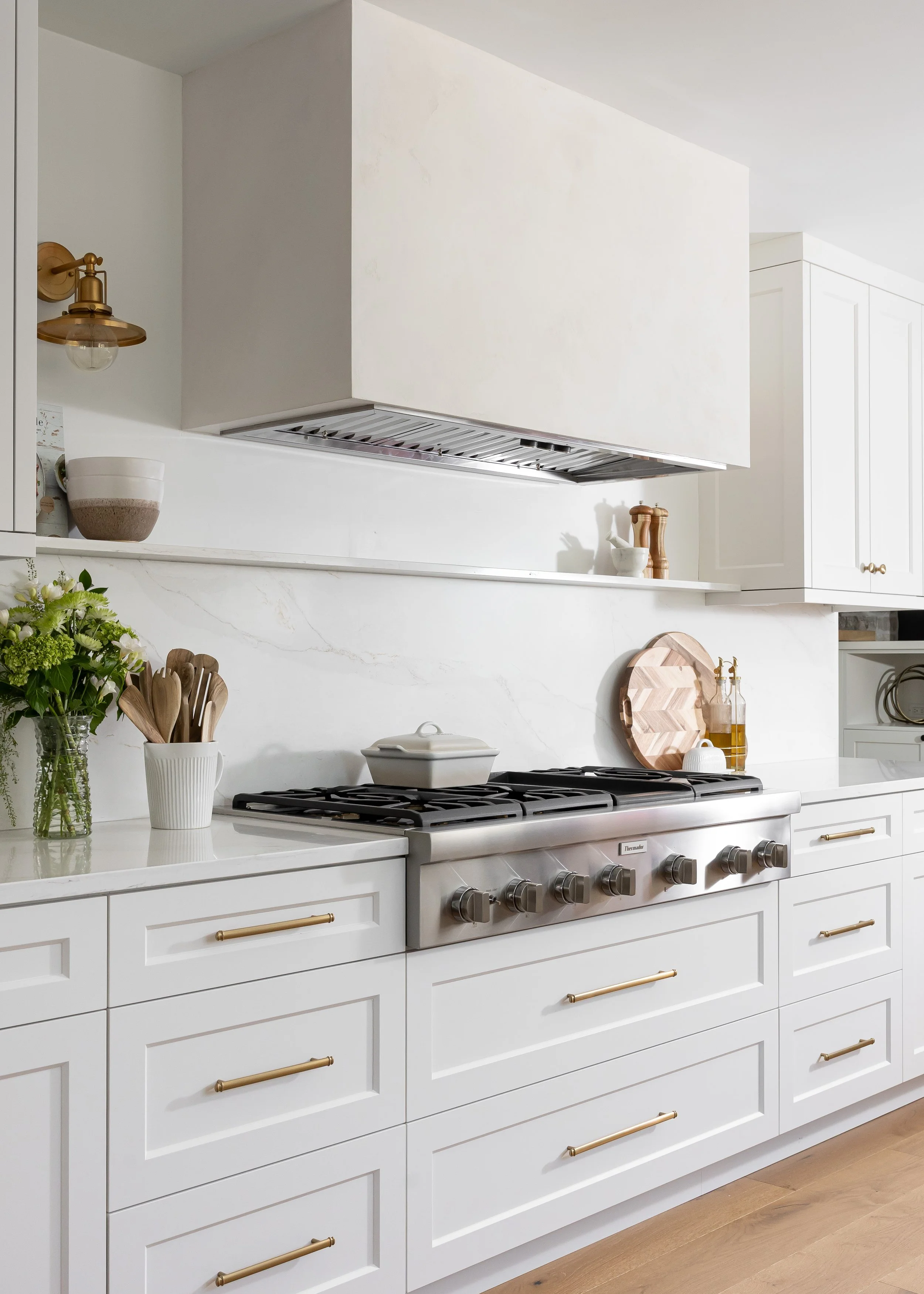 Studio 7 Designs Custom Home Interior Design of A modern white kitchen with cabinets, a stove, a vent hood, and decorative items including a potted plant, bowls, kitchen utensils, and bottles on the countertop.