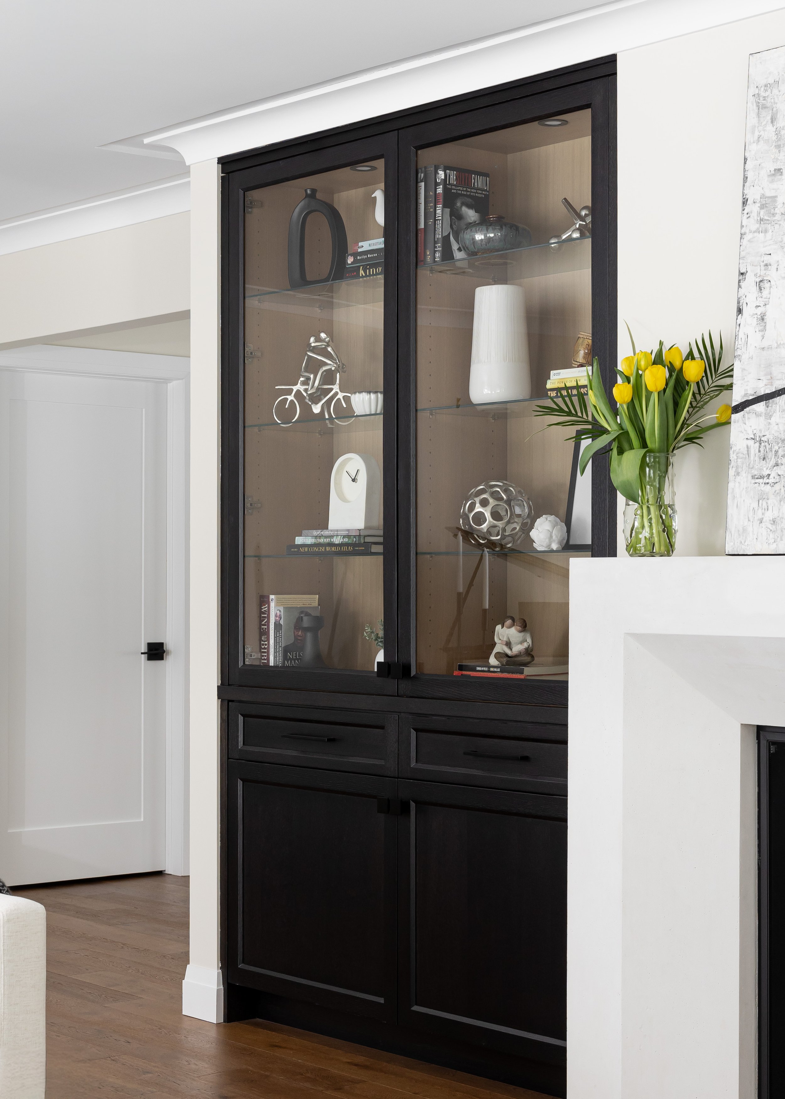 Stylish built-in black display cabinet in a modern living room designed by Studio 7 Designs. The cabinet features glass doors showcasing curated decorative objects, books, and vases for aesthetic storage. Below, concealed cabinetry offers practical s