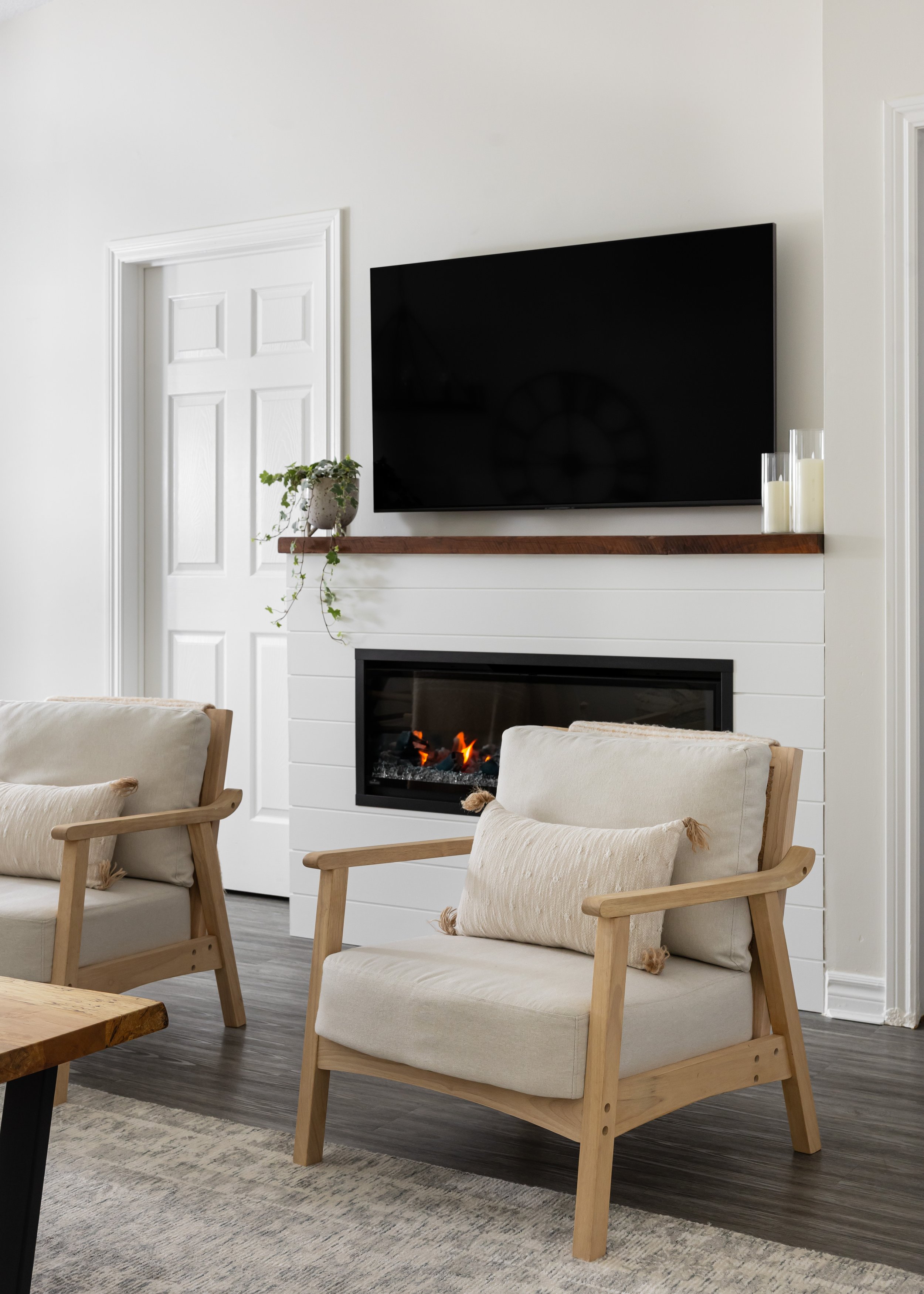 Transitional living room featuring a white shiplap fireplace with a rustic wood mantel, wall-mounted TV, and cozy armchairs—contemporary interior styling by Studio 7 Designs.