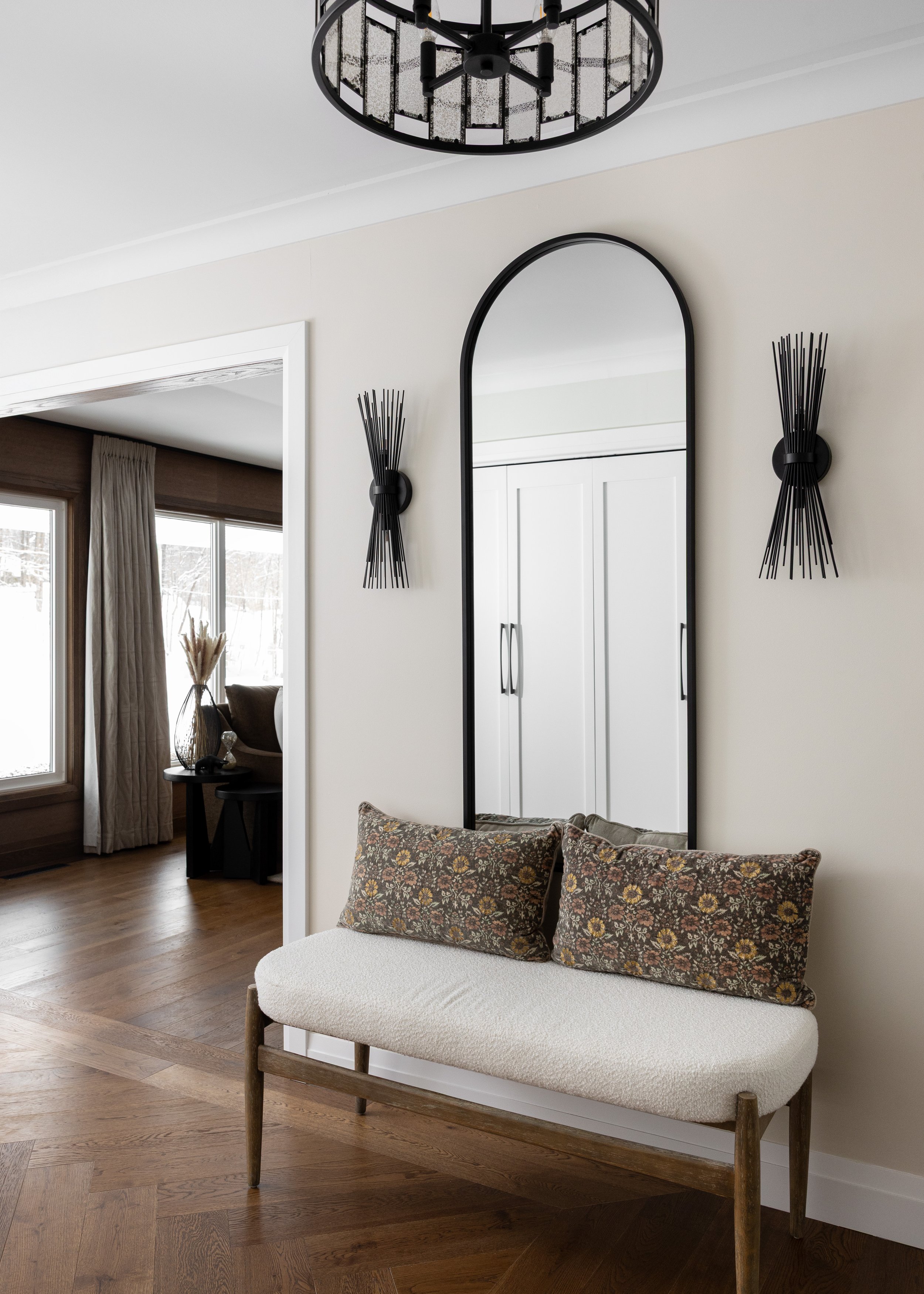 Chic entryway by Studio 7 Designs featuring a soft boucle bench with vintage floral cushions, flanked by sculptural black wall sconces and a tall arched mirror. Warm herringbone wood floors and black iron ceiling lighting add contrast to the clean, m
