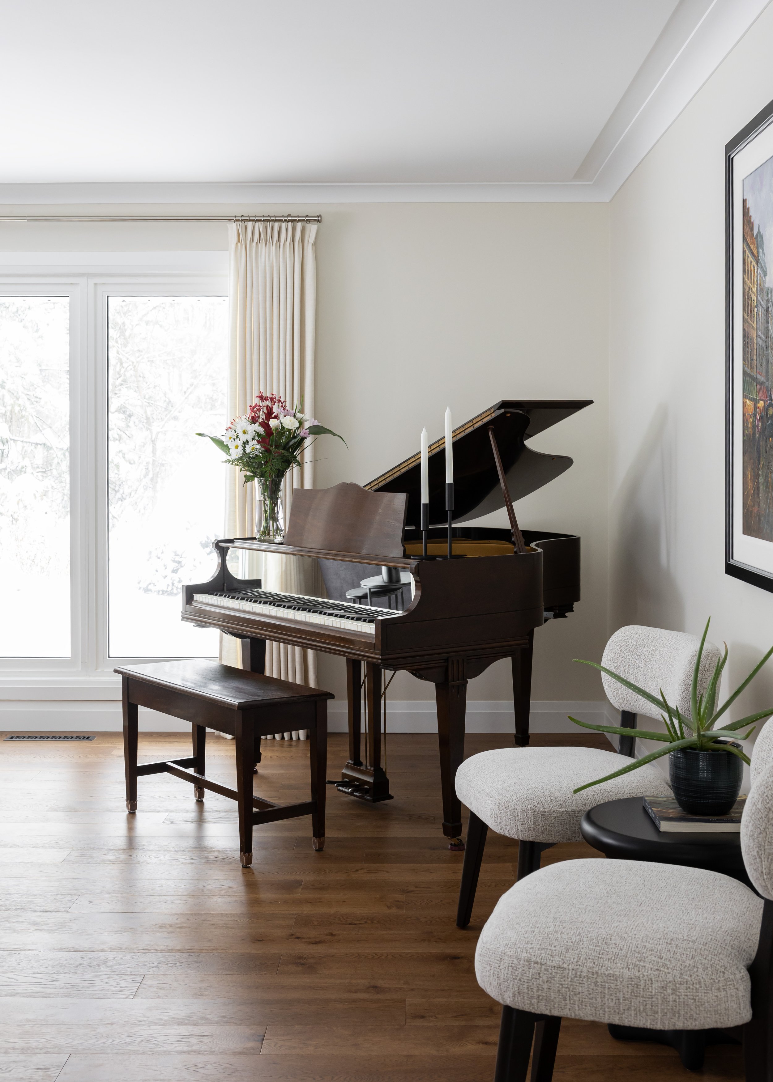 Elegant living room by Studio 7 Designs featuring a rich wood baby grand piano paired with a matching bench, styled with fresh florals. Soft boucle accent chairs and minimalist décor create a serene corner against wide plank wood floors and creamy dr