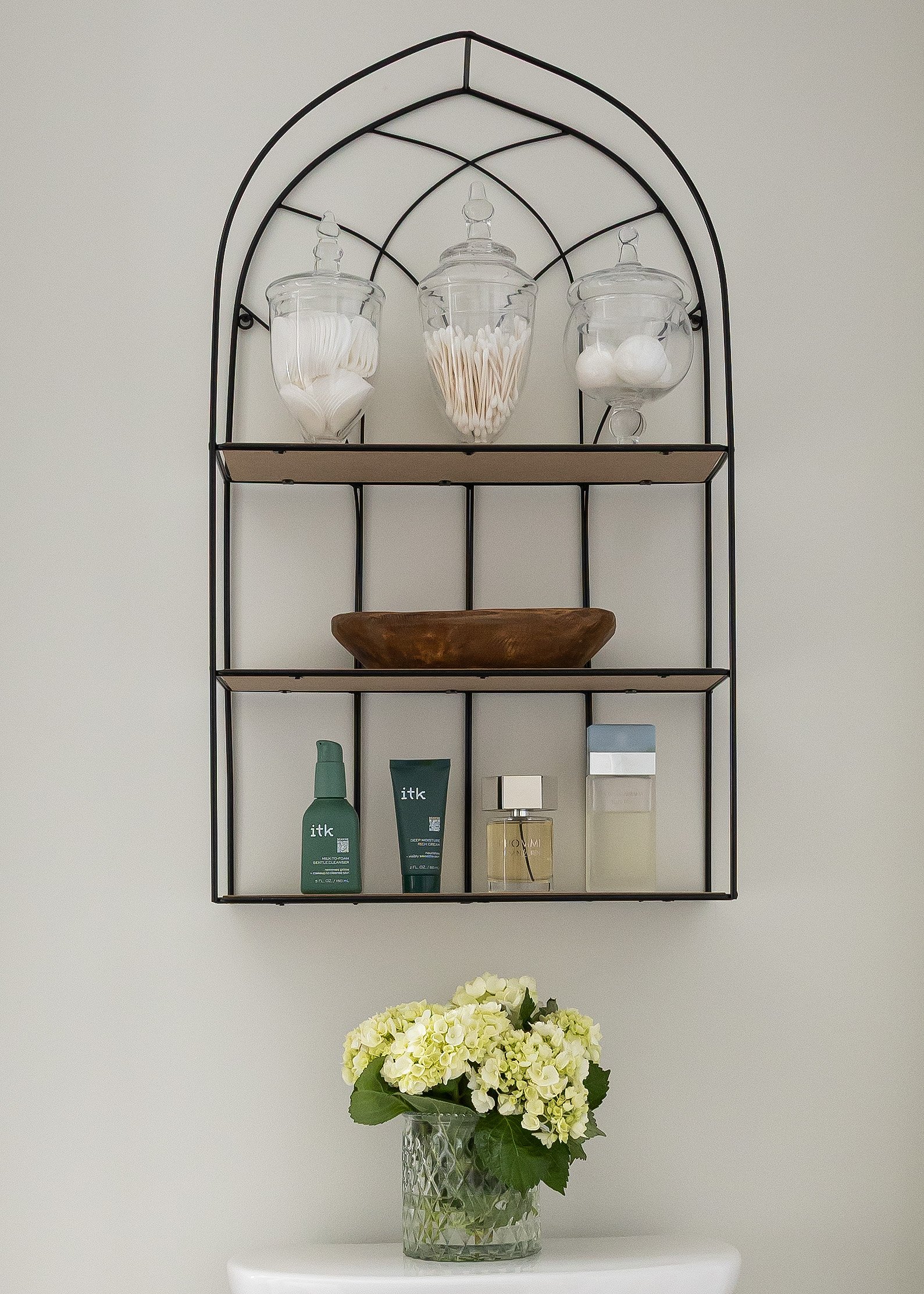 Bathroom wall shelf styling by Studio 7 Designs, featuring a black arched metal shelf with glass apothecary jars filled with cotton rounds, swabs, and balls, a wooden bowl, skincare essentials, designer fragrances, and a crystal vase with fresh hydra