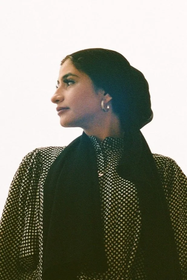 A woman with dark hair tied back, wearing a polka dot blouse, hoop earrings, and a black scarf, looking to the side against a plain white background.