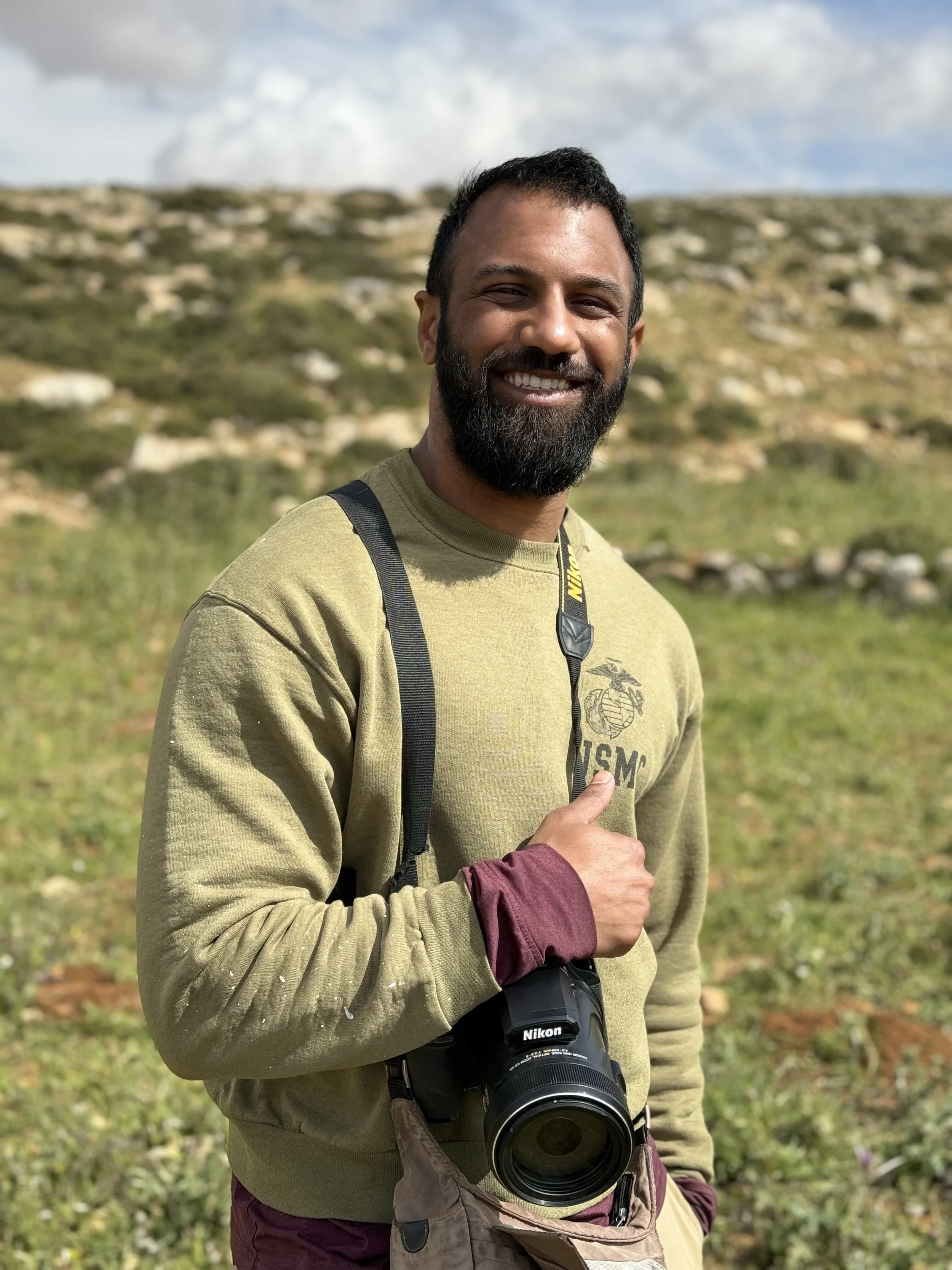 A smiling man with a beard in an olive green sweatshirt holding a Nikon camera outdoors in a grassy field with rocks and a cloudy sky in the background.