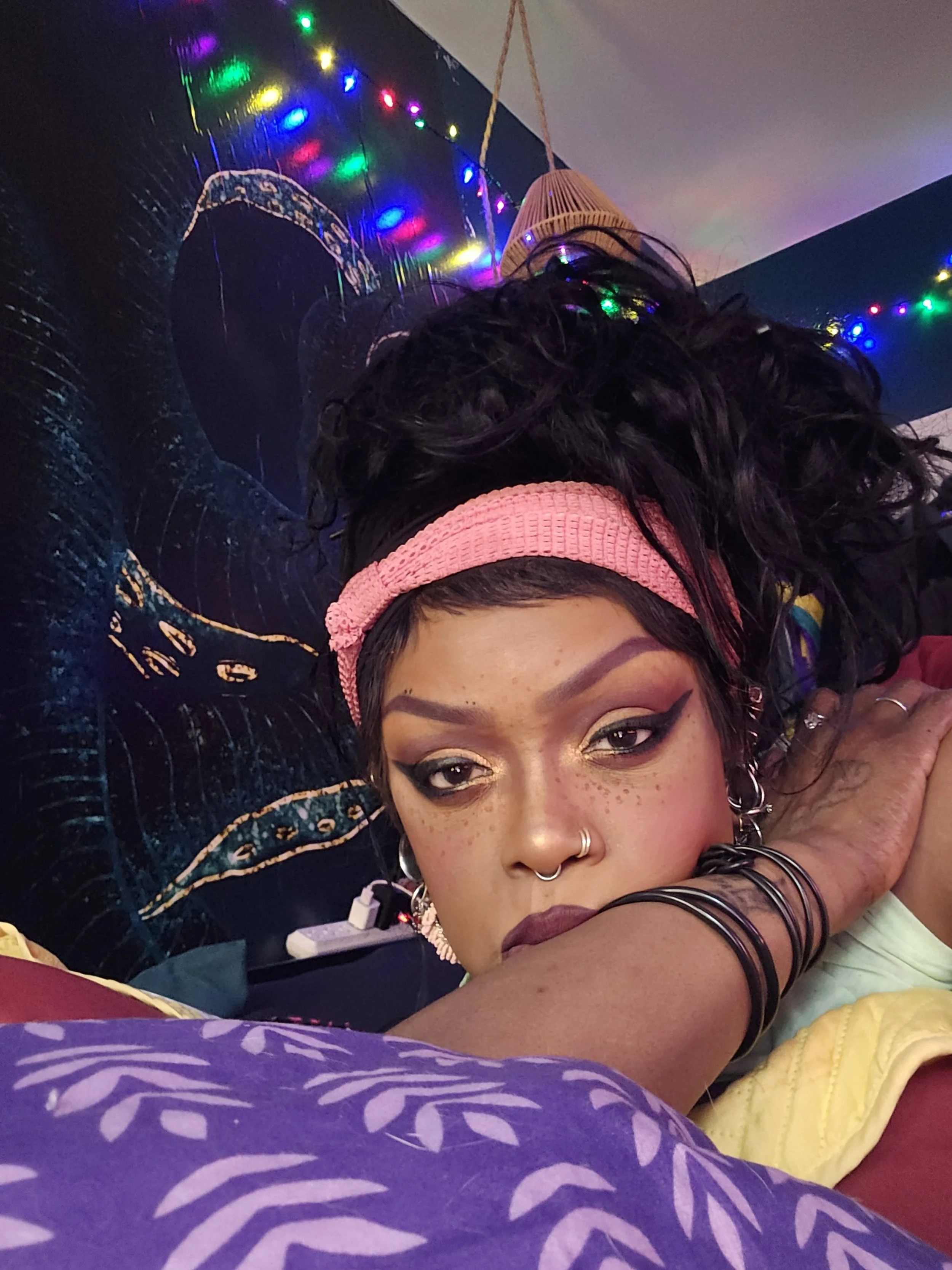 A woman with dark curly hair wearing a pink headband, gold and dark eye makeup, and multiple earrings. She rests her face on an arm in a colorful, dimly lit room decorated with multicolored string lights and a tapestry with a tiger design.