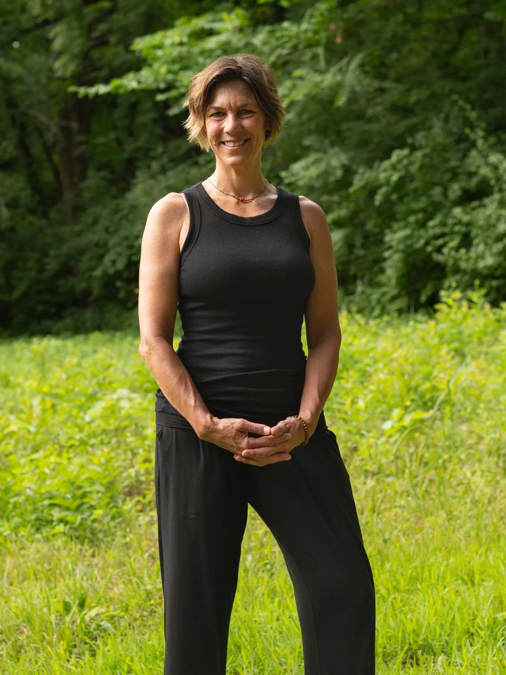 A woman standing outdoors in a green, wooded area, smiling and wearing a sleeveless black top and black pants.