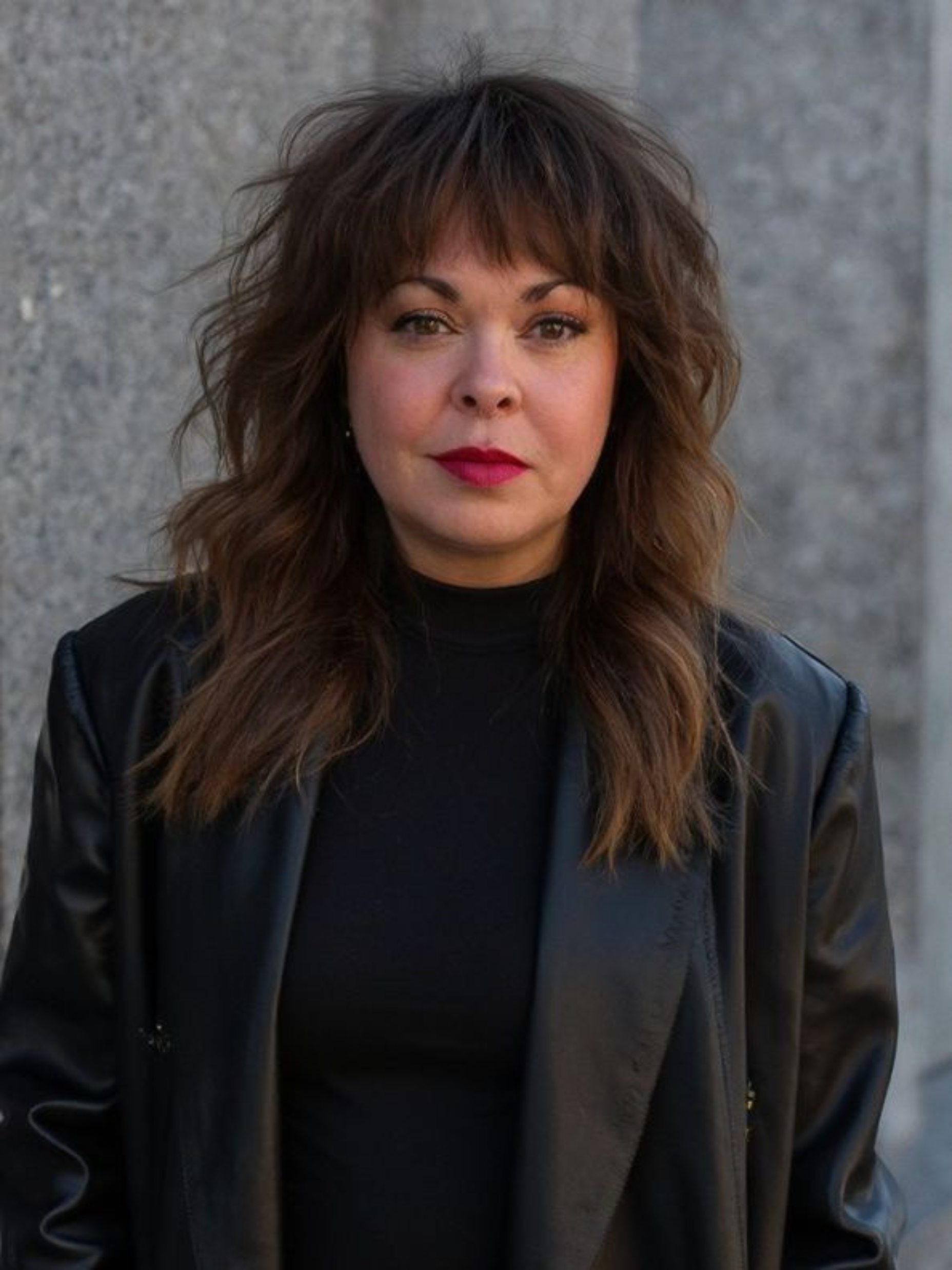 Close-up image of a woman with shoulder-length brown hair and bangs, wearing a black top and black leather jacket, standing in front of a gray stone wall.