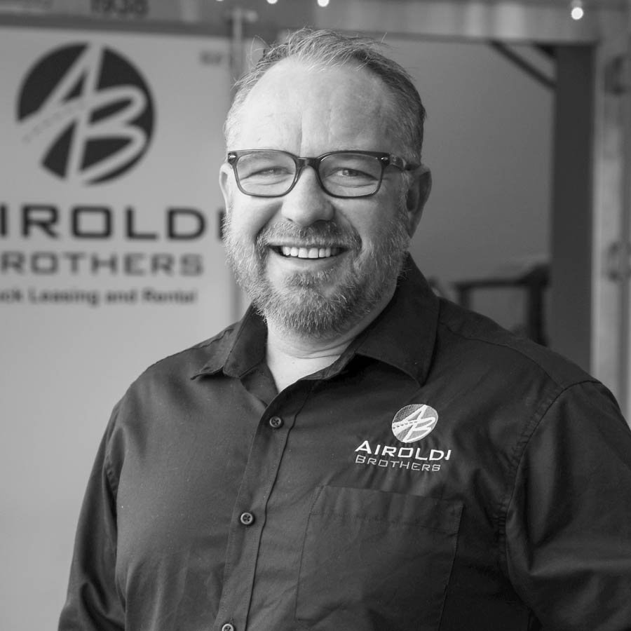 A smiling man with glasses and a beard, wearing a black shirt with a logo that reads 'AROLDI BROTHERS'. In the background, there is a sign that also reads 'AIOLDI BROTHERS'.