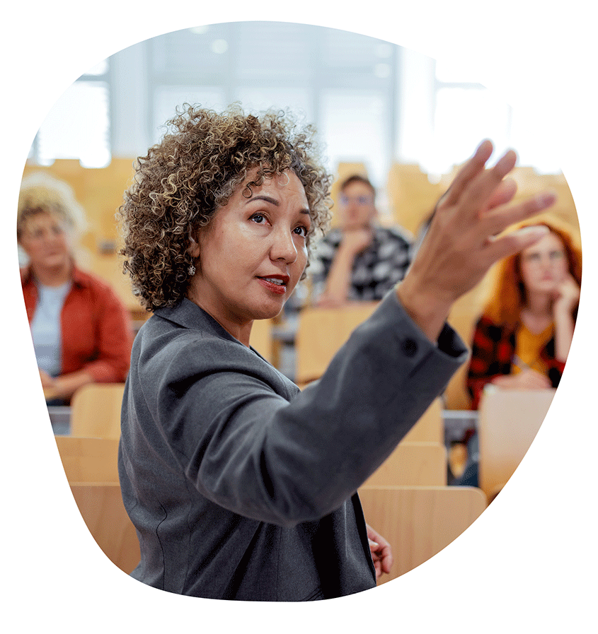 A teacher is pointing to the screen behind her in an auditorium-style college classroom.