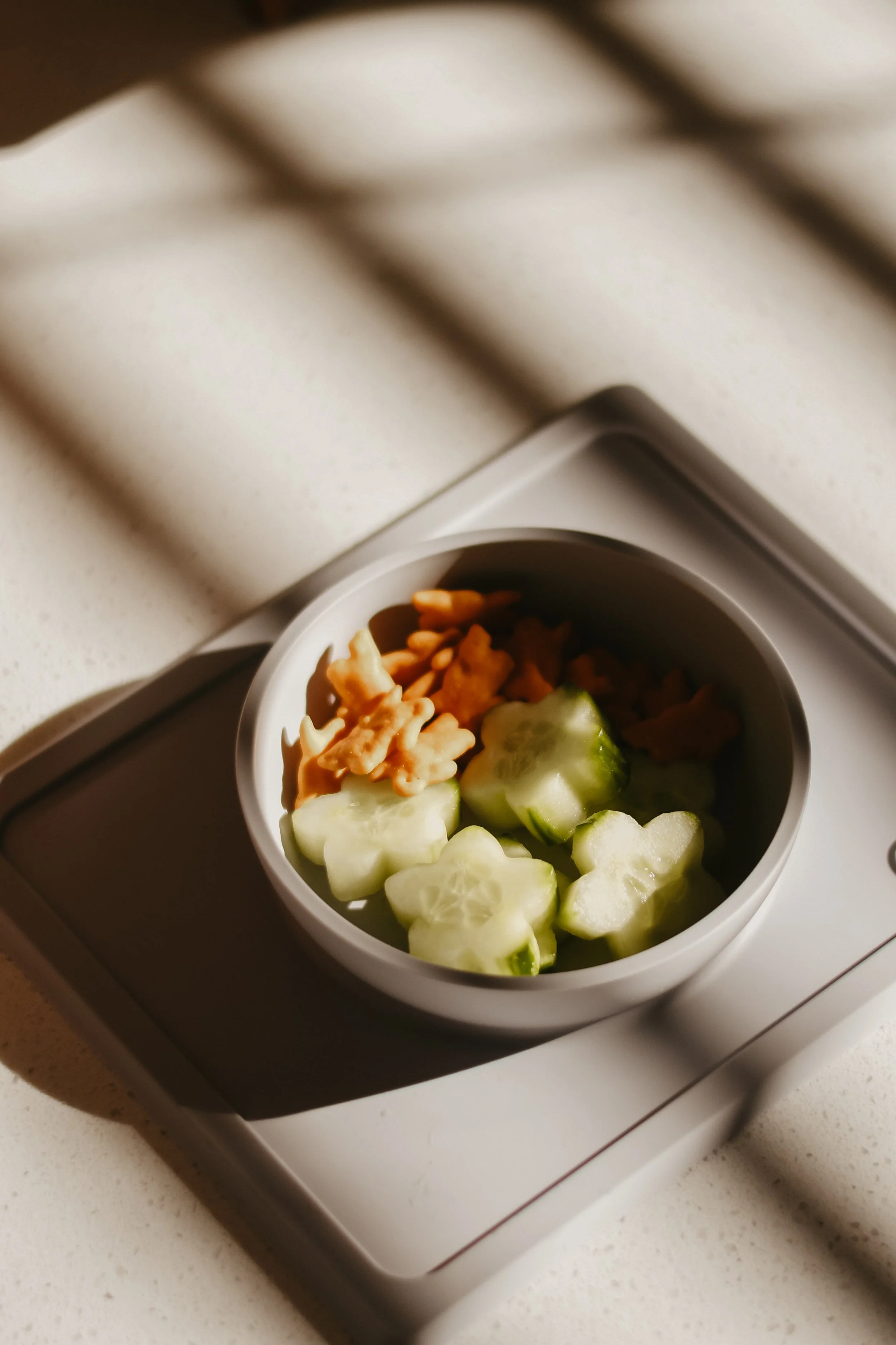Cucumber slices and peanut butter bites in a white bowl on a kitchen scale with shadows cast by window blinds.