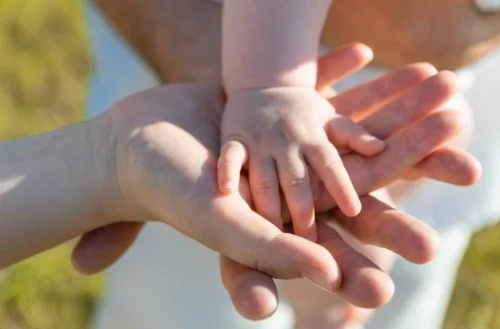 An adult hand holding a baby's hand outdoors with grass and sky in the background. Nurturing pediatrics
