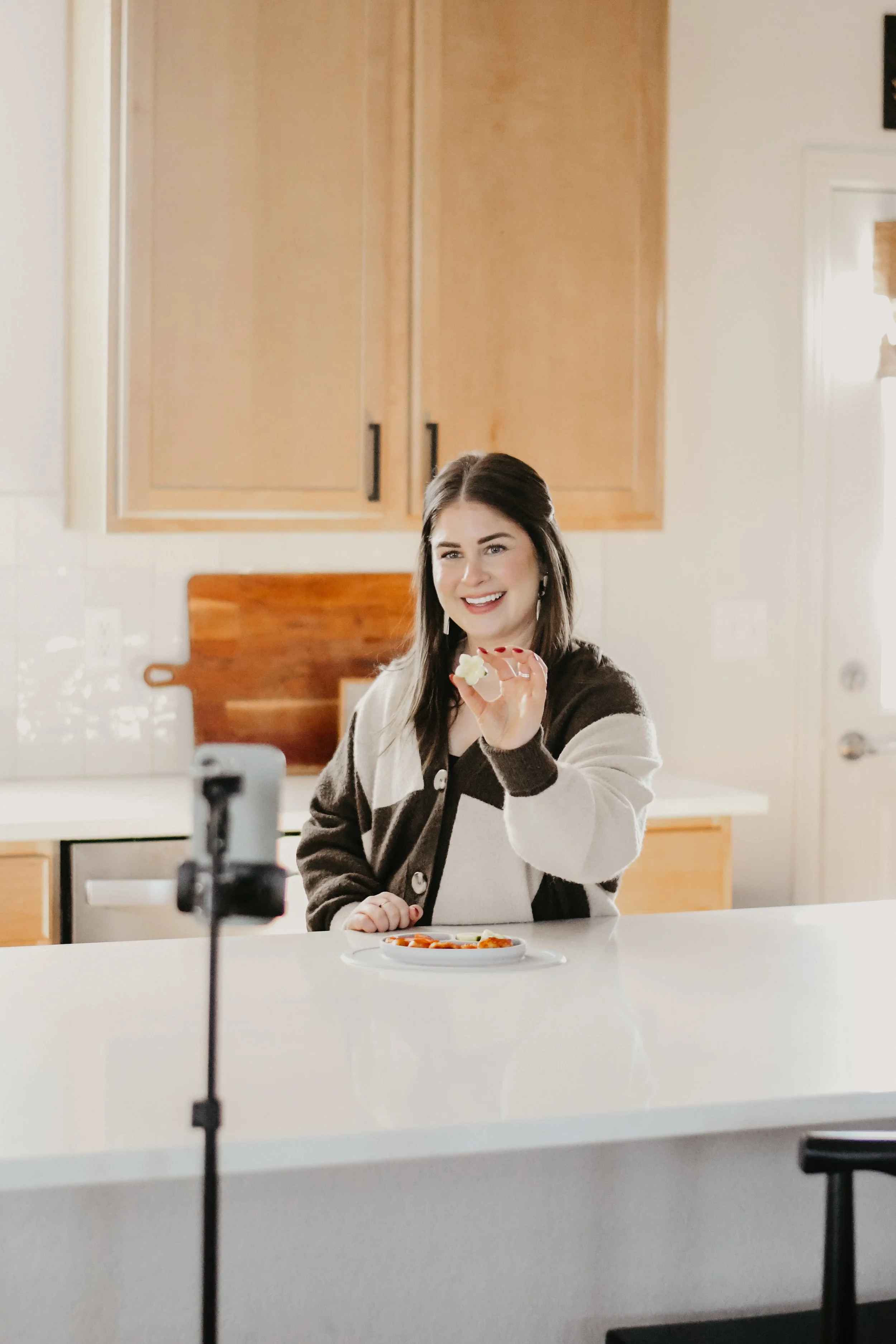 A woman with dark hair smiling and showing a slice of pizza, sitting at a white kitchen counter with a plate of food in front of her. She is holding the pizza towards a camera, and a camera setup on a tripod is visible in the foreground.
