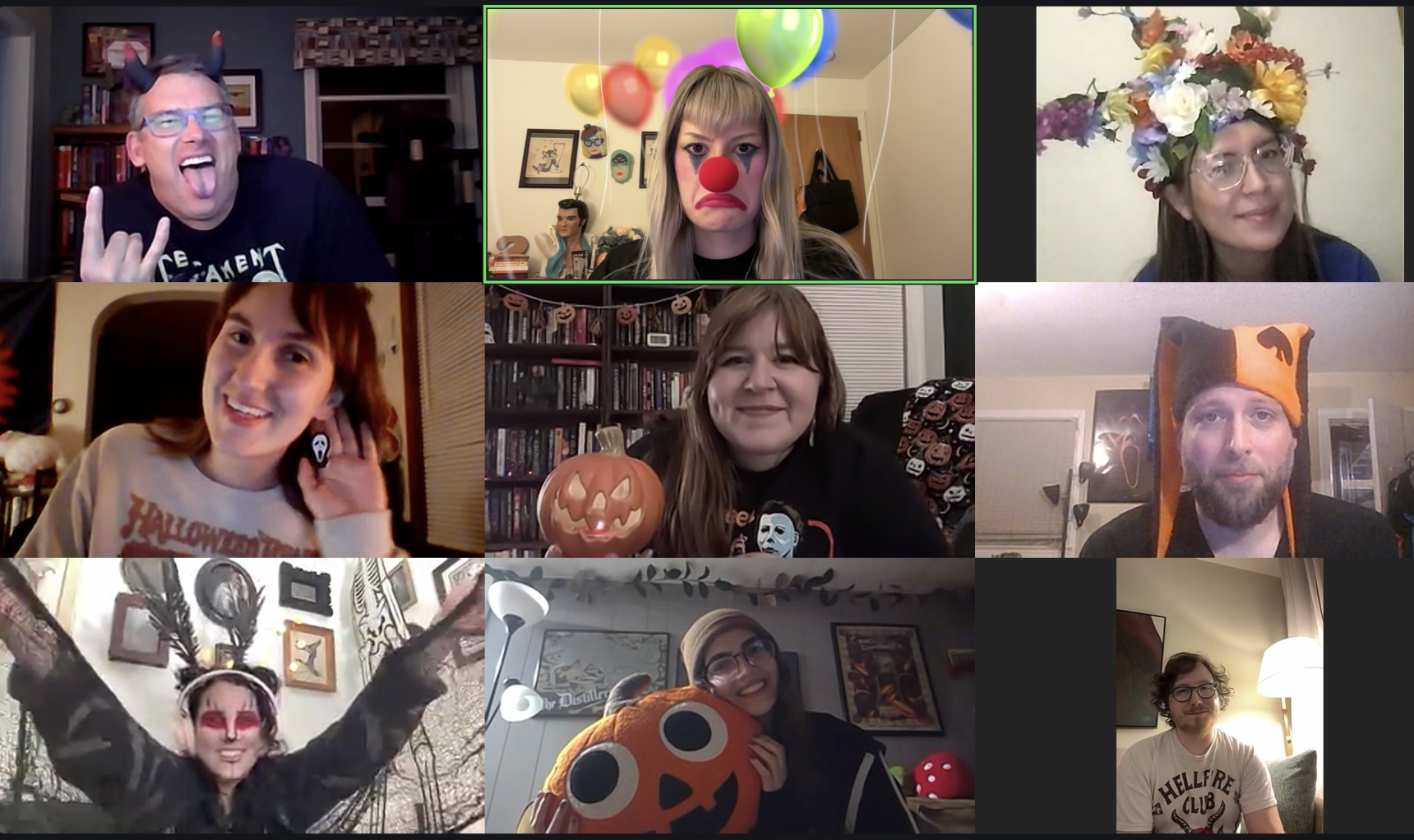 A group of nine people participating in a virtual Halloween-themed video call, with some wearing costumes and accessories like clown makeup, a witch's hat, a pumpkin, and Halloween-themed face paint.