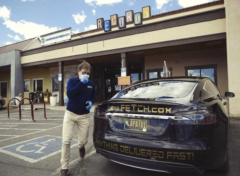 Person in protective gear standing by a parked black car with 'FETCH' website and delivery slogan, in front of a storefront with a 'RECORDS' sign.